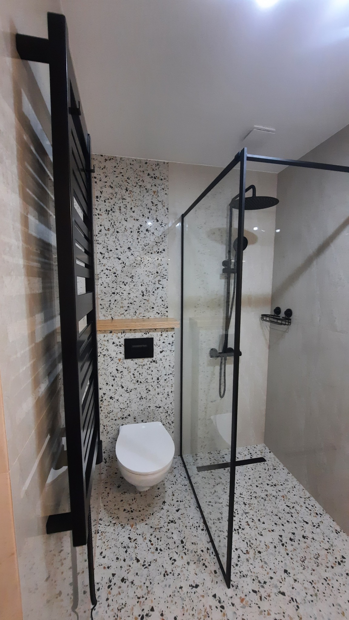 Nowoczesna łazienka z kabiną typu walk-in, wykończona płytkami lastryko. Czarny grzejnik drabinkowy i armatura podkreślają minimalistyczny design. Widoczna toaleta i półka.