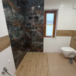 Wykończona łazienka z kabiną prysznicową bez brodzika, wyłożoną płytkami imitującymi marmur w odcieniach czerni i brązu, drewnopodobną podłogą, białą ceramiką i oknem z zasłoniętą szybą.