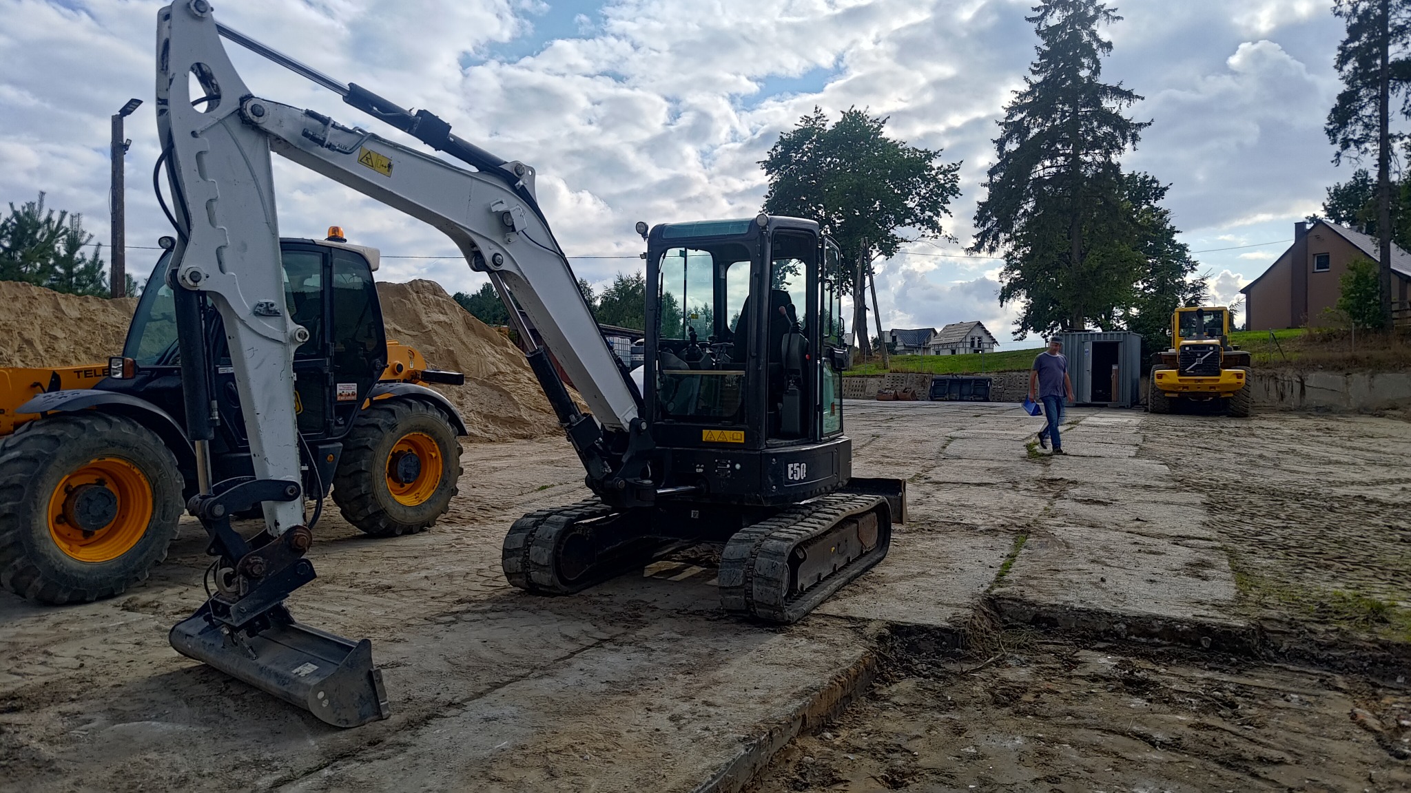 Kompaktowa koparka gąsienicowa E50 na betonowej płycie, obok ładowarka teleskopowa i spora pryzma piasku, w tle budynki mieszkalne i idący mężczyzna.
