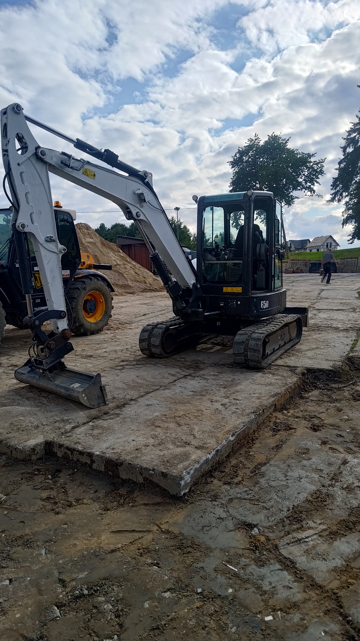 Kompaktowa koparka E50 na betonowych płytach w Żukowie, obok widoczna ładowarka i sterta ziemi, w tle budynki mieszkalne i pochmurne niebo.