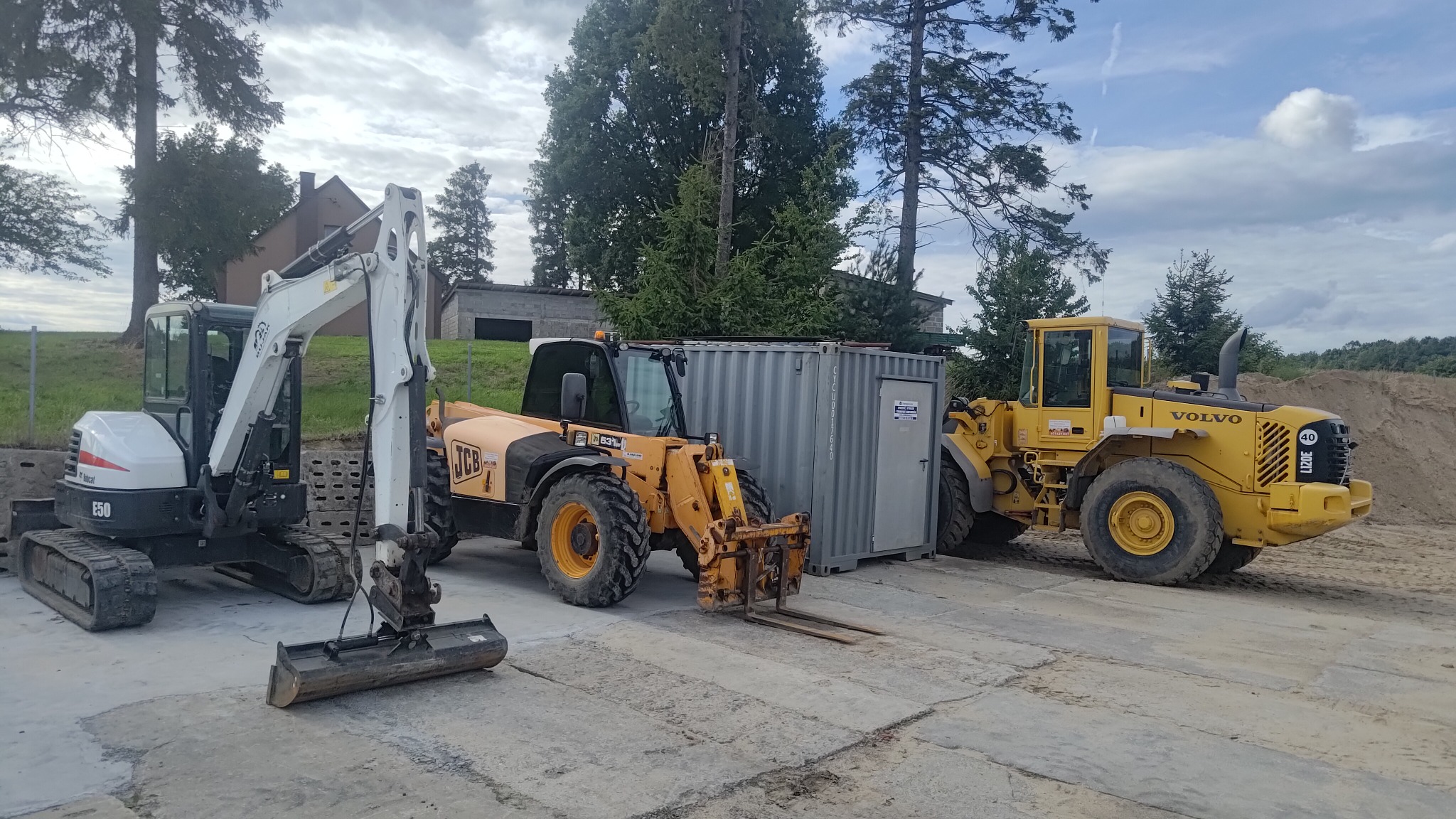Park maszyn budowlanych: biała minikoparka Bobcat E50 z łyżką, żółty wózek widłowy JCB 531-70 i żółta ładowarka kołowa Volvo L120E, stojące obok szarego kontenera na betonowym placu, w tle drzewa...