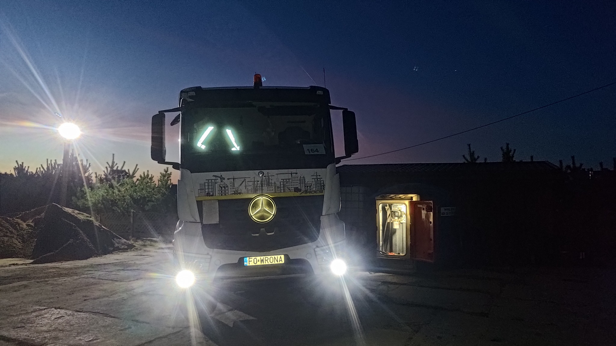 Ciężarówka z logo firmy budowlanej zaparkowana na placu budowy o zmierzchu, obok otwartej budki z oświetleniem wewnątrz.