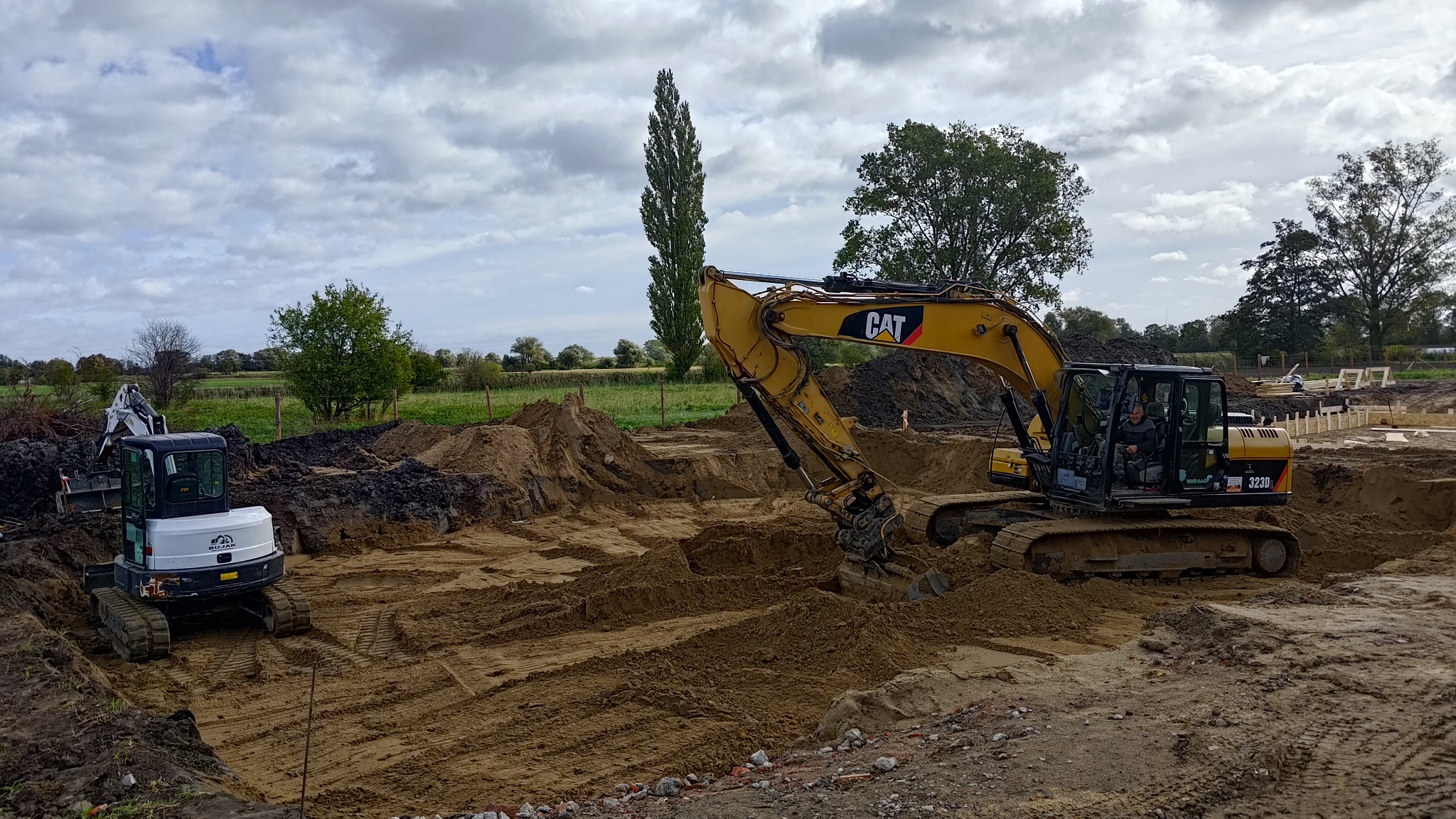 Dwie koparki, żółta CAT 323D i biała BUDAJ, pracują na placu budowy, widoczne wykopane rowy i pryzmy ziemi, w tle drzewa i pochmurne niebo.
