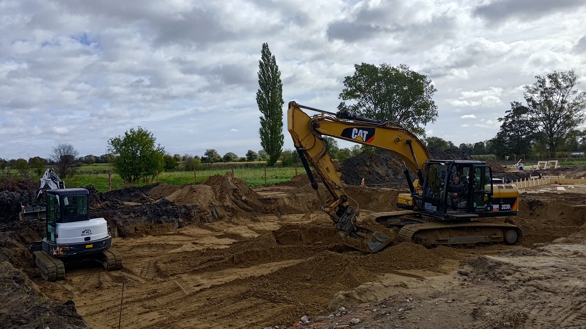 Żółta koparka CAT 323D w wykopie, operator w kabinie, obok mniejsza biała koparka, widoczne drewniane szalunki, ziemia i fragment krajobrazu z drzewami pod pochmurnym niebem.