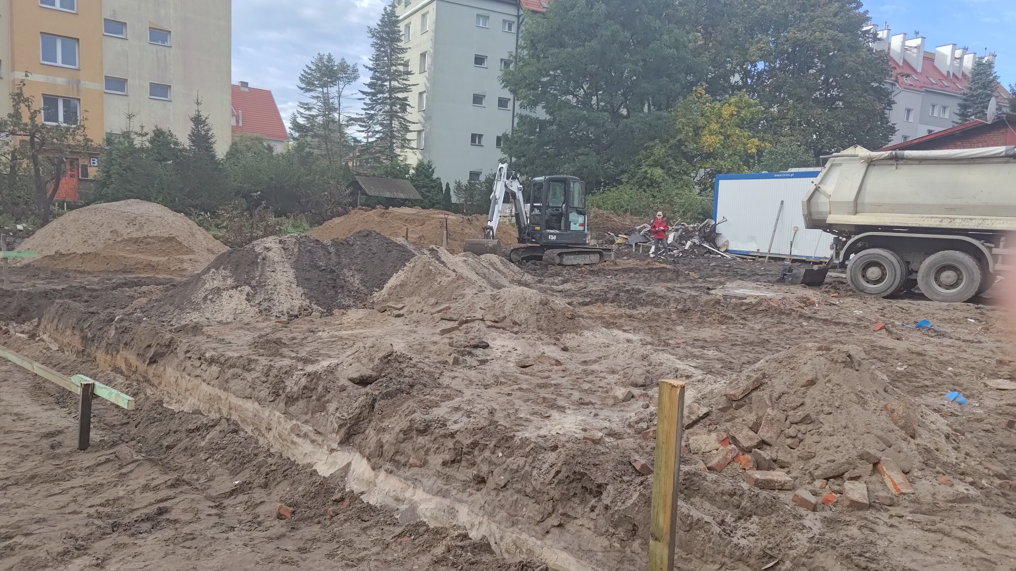 Wykop pod fundamenty z widocznym wykopanym rowem, stertami ziemi, mini koparką Bobcat E50, ciężarówką i pracownikiem w czerwonej kurtce w tle.