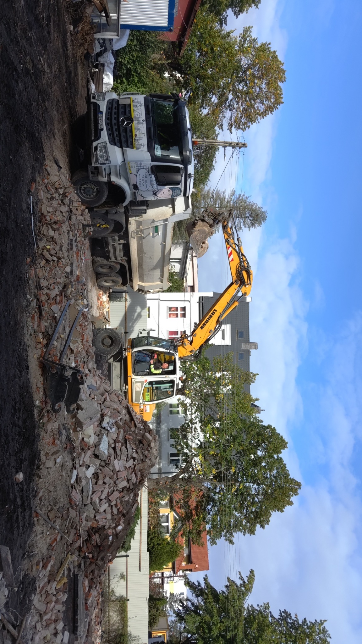 Żółta koparka Liebherr załadowuje gruz ceglany do białej ciężarówki Mercedes-Benz na tle budynków i drzew, widoczne logo firmy na kabinie ciężarówki.