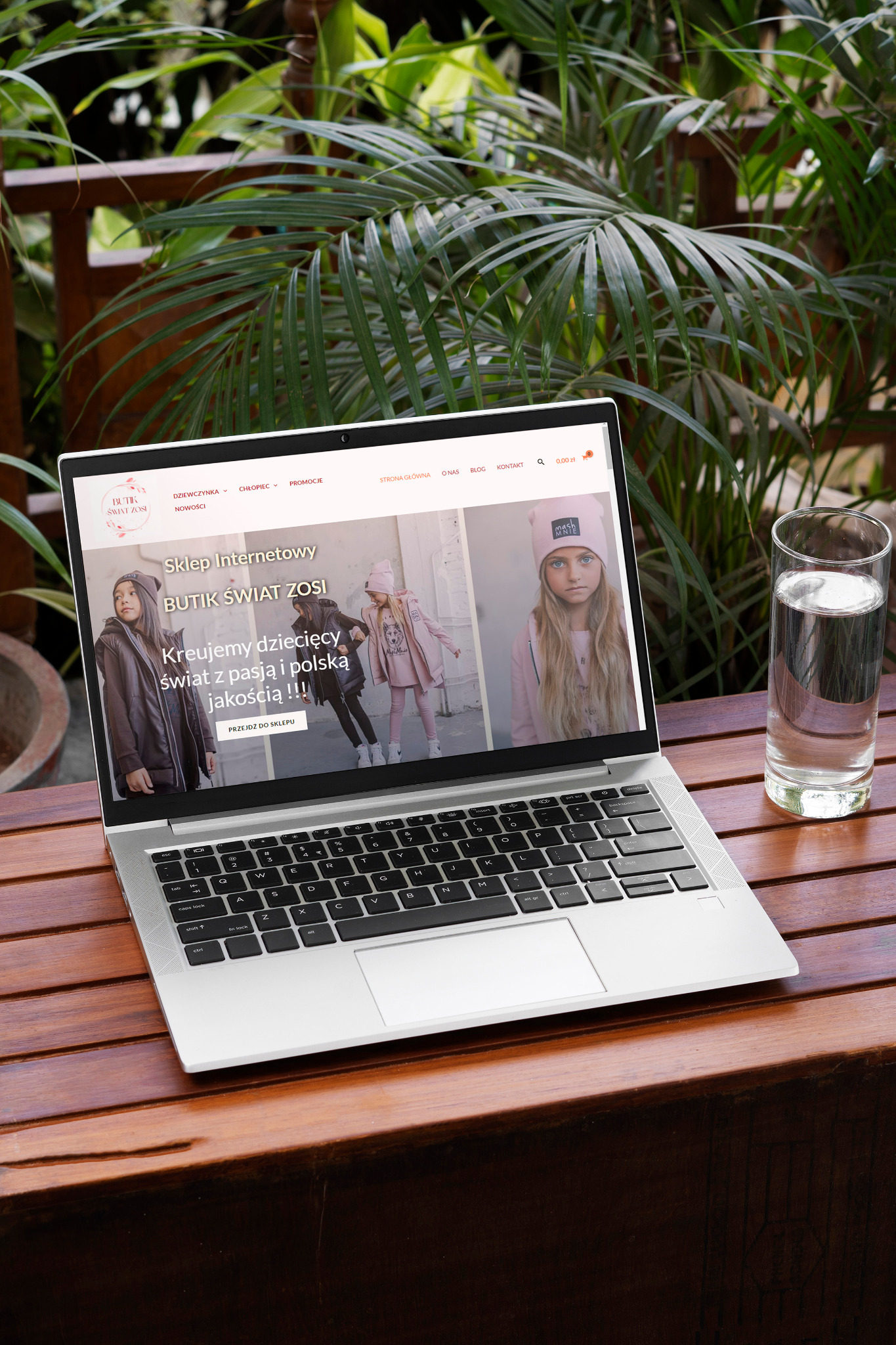 Laptop wyświetla stronę internetową sklepu z odzieżą dziecięcą 'Butik Świat Zosi' na drewnianym stole, obok szklanka wody i tropikalne rośliny w tle.