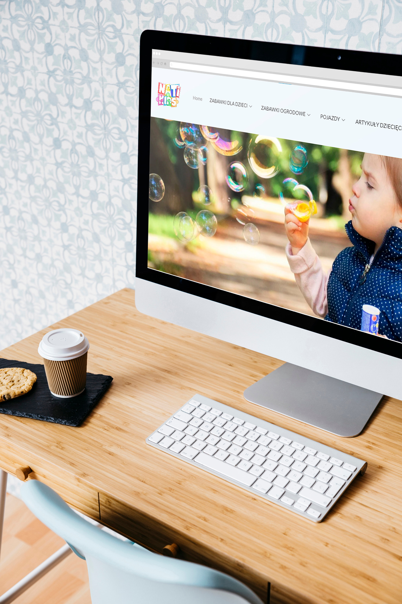 Ekran monitora wyświetla stronę internetową sklepu z zabawkami dla dzieci; na biurku obok monitora leży ciastko, kawa i klawiatura.