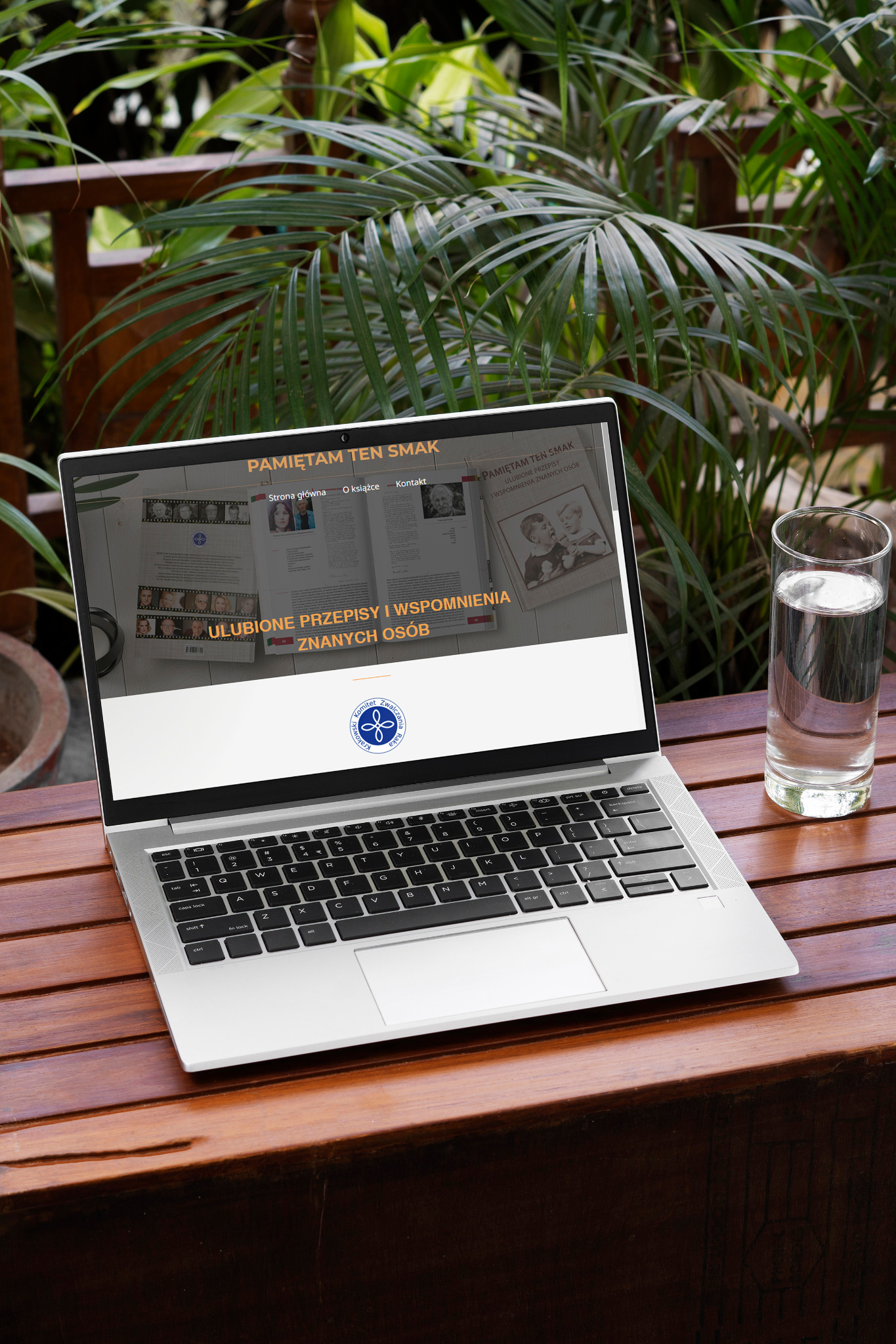 Laptop wyświetlający stronę internetową z tytułem 'Pamiętam ten smak' na drewnianym stole, obok szklanka wody, w tle roślinność.