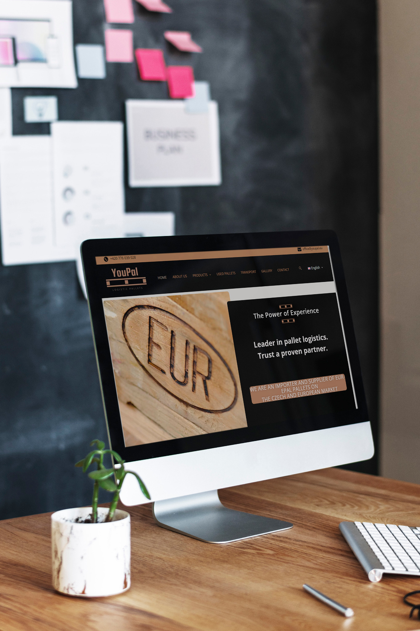Monitor wyświetla stronę internetową firmy YouPal, specjalizującej się w logistyce palet EUR, z widocznym logo i hasłem 'The Power of Experience', umieszczony na drewnianym biurku obok klawiatury...