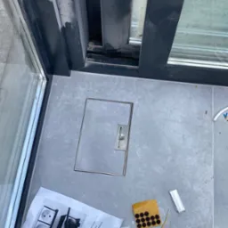 Szare płytki podłogowe z odpływem liniowym przy oknie przesuwnym, widoczne akcesoria montażowe i instrukcja montażu gniazdek elektrycznych.