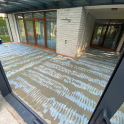 Przygotowanie podłoża pod panele w przeszklonej werandzie: wylewka z widoczną siatką wzmacniającą, duże okna z brązowymi ramami, szara cegła elewacyjna.