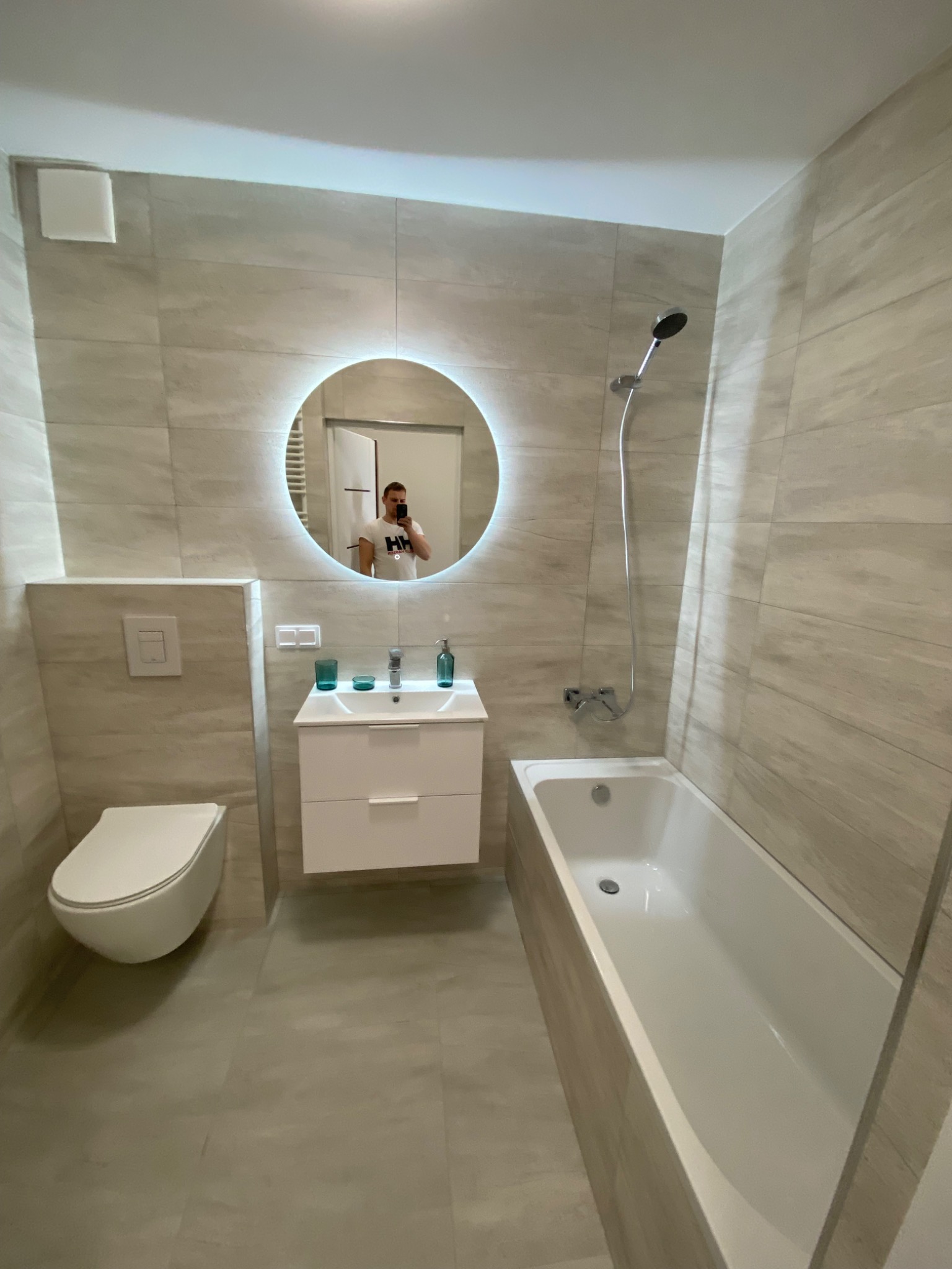 Nowoczesna łazienka z beżowymi płytkami imitującymi drewno, okrągłe lustro z podświetleniem LED, biała umywalka z szafką i bateria, wisząca toaleta oraz biała wanna z chromowaną armaturą prysznicową.