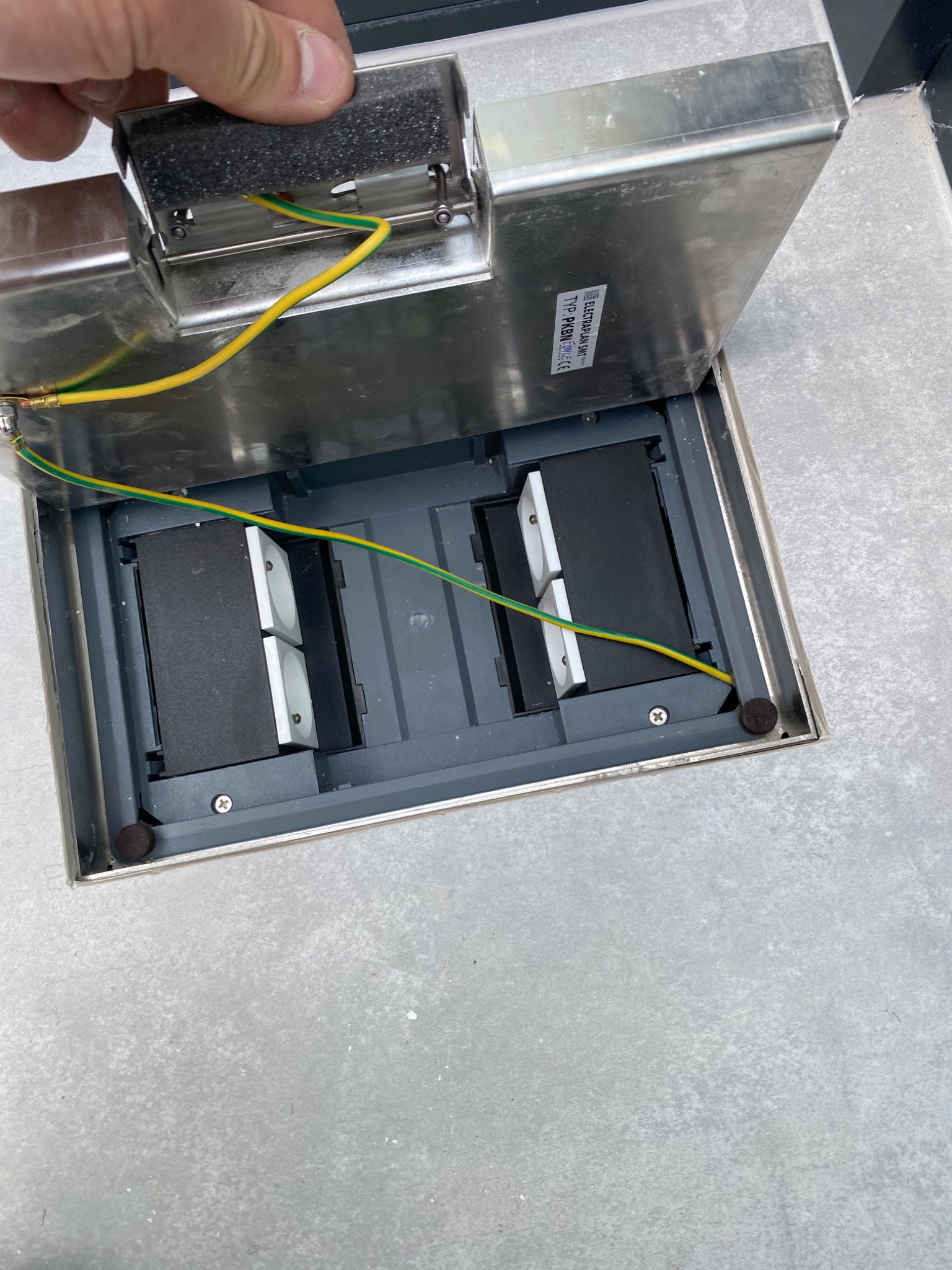 Otwarta, metalowa puszka podłogowa z dwoma gniazdami elektrycznymi, widoczne przewody zasilające i uziemienie.