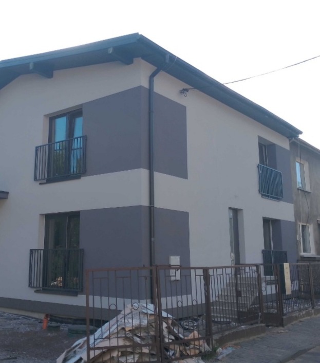 Dwukondygnacyjny dom z jasnoszarą elewacją z grafitowymi pasami, balkonami z metalowymi barierkami i metalowym ogrodzeniem, stos materiałów budowlanych przed ogrodzeniem.