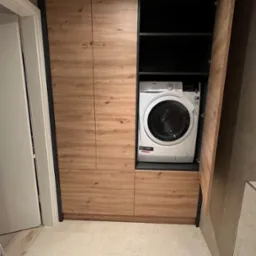 Zabudowa pralki w łazience z drewnopodobnymi frontami szafek i czarnymi elementami konstrukcyjnymi, widoczny fragment toalety i drabiny.