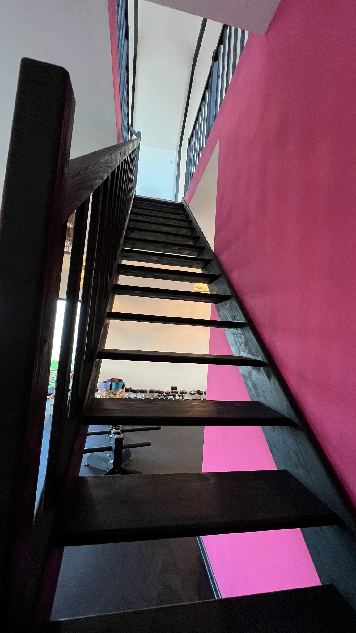 Wysokie, ciemne schody drewniane prowadzące w górę, z różową ścianą po prawej stronie i widocznym wyposażeniem siłowni na dole.
