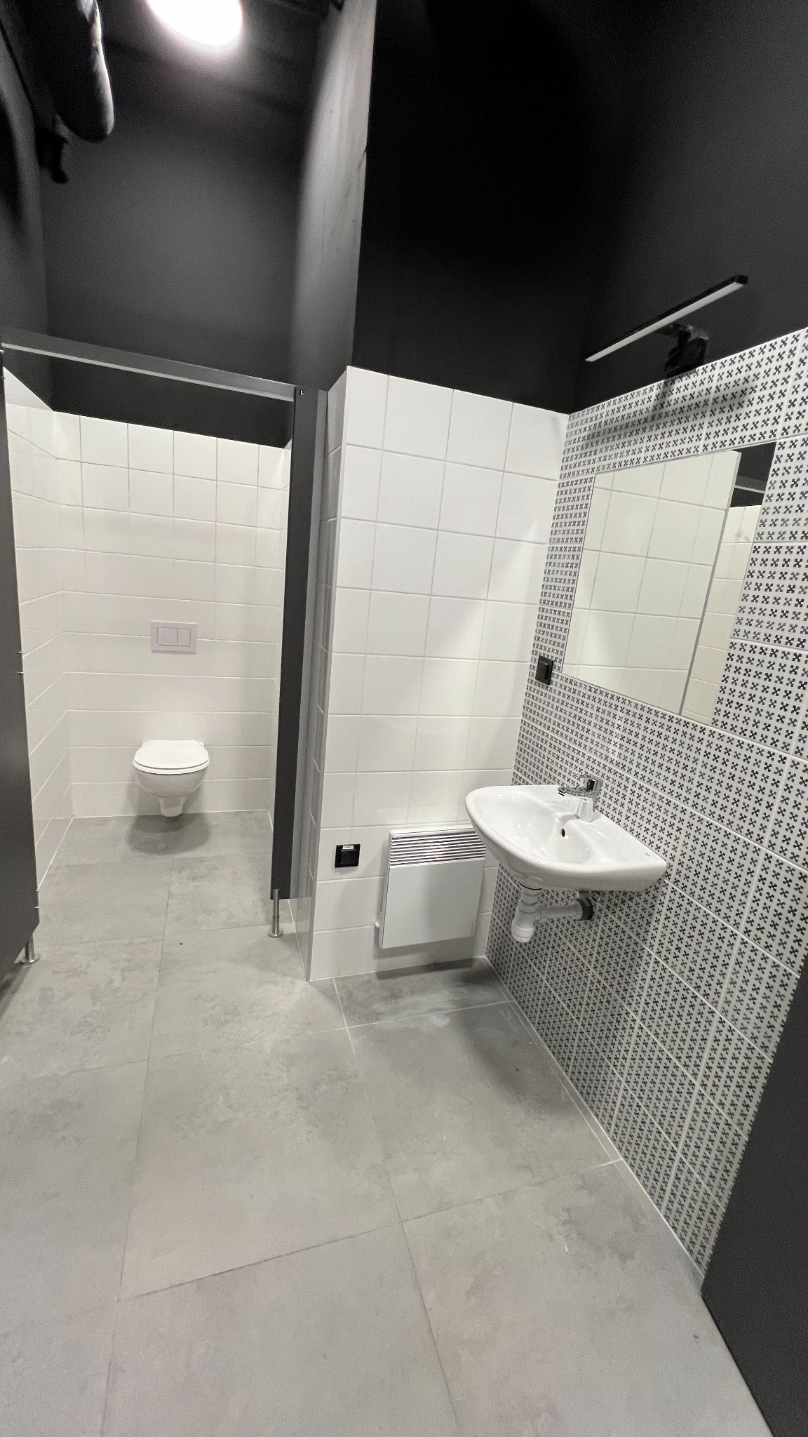 Nowoczesna łazienka w biurze z umywalką, toaletą i szarą podłogą z płytek, białe płytki na ścianach częściowo z wzorem, czarny sufit