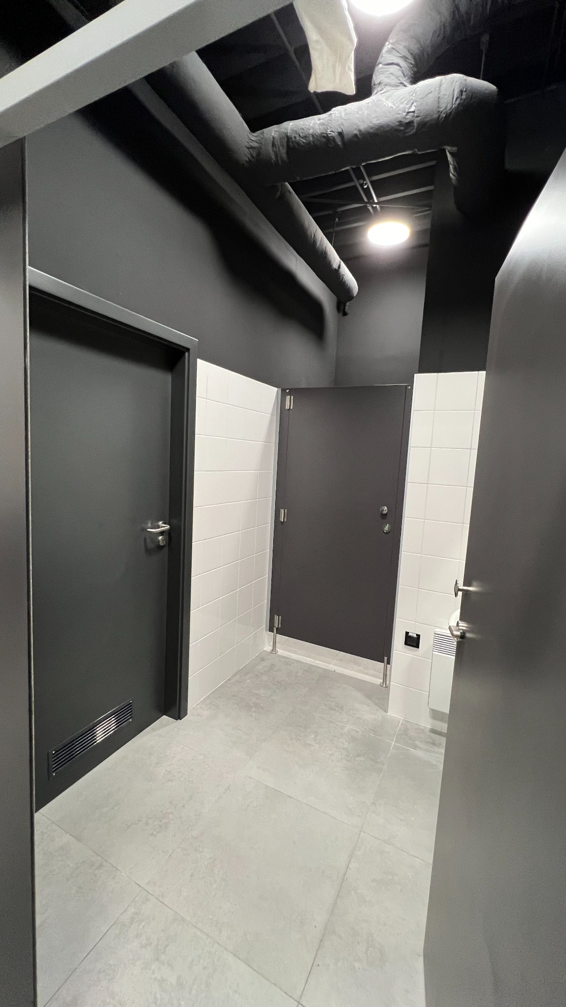 Wnętrze nowoczesnej łazienki publicznej z trzema kabinami oddzielonymi białymi płytkami, widoczne czarne rury pod sufitem i szara podłoga z płytek.