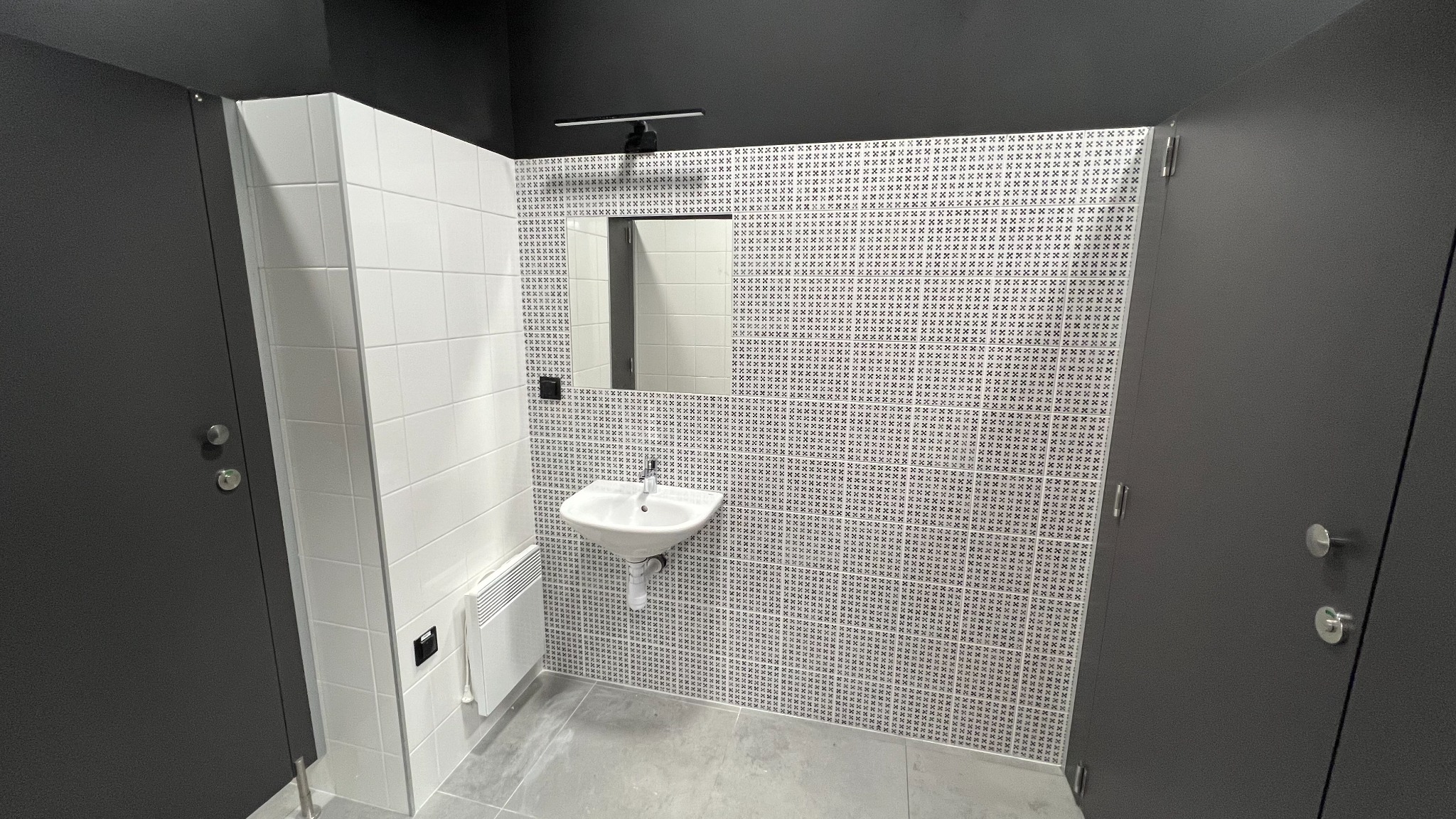 Nowoczesna łazienka publiczna z kabinami toaletowymi w kolorze antracytowym, białą umywalką ścienną, lustrem z oświetleniem i płytkami w geometryczny wzór na ścianie.