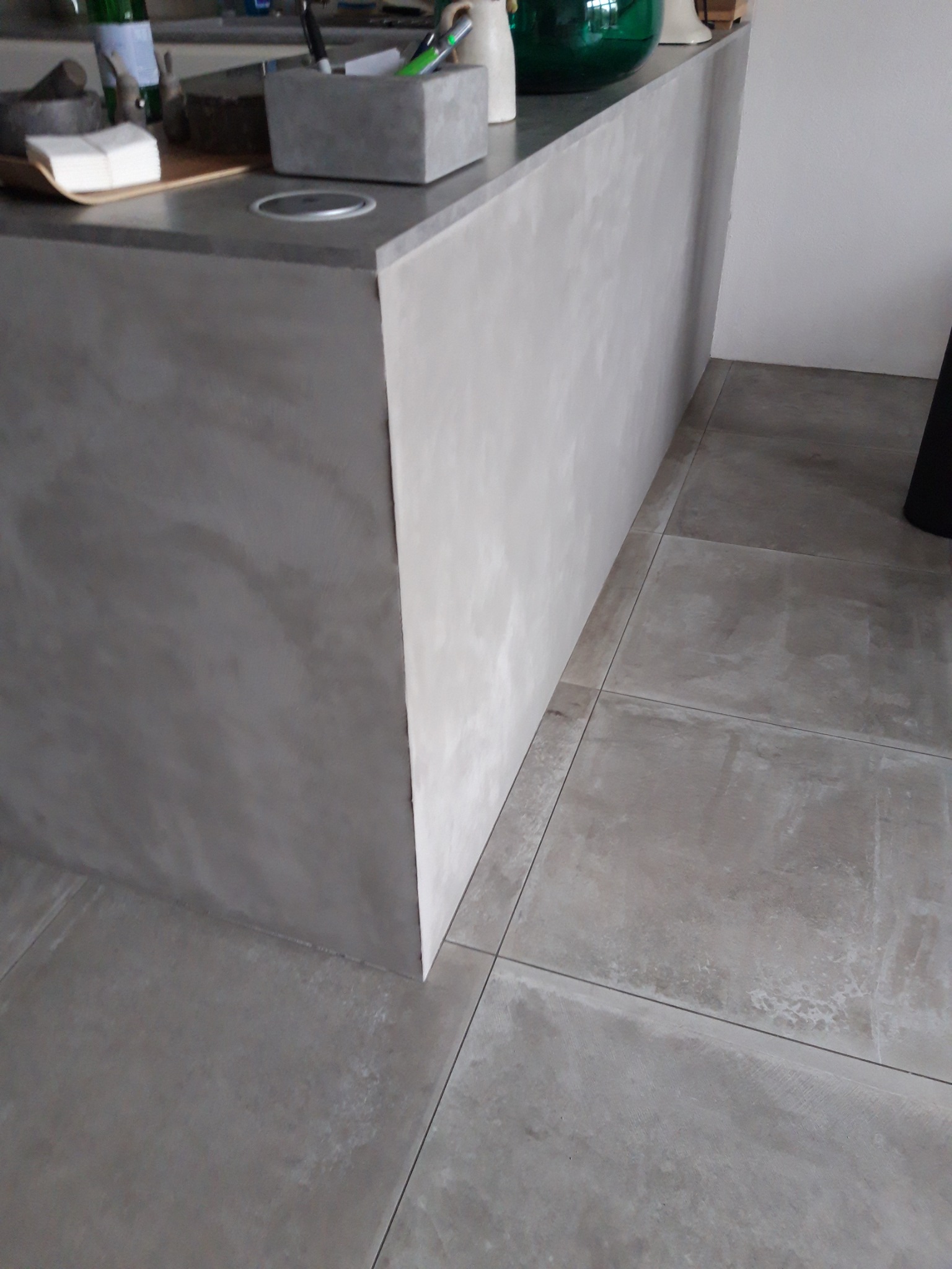 Szara wyspa kuchenna wykończona mikrocementem, z widocznym blatem i bokiem, oraz pasującymi płytkami podłogowymi.