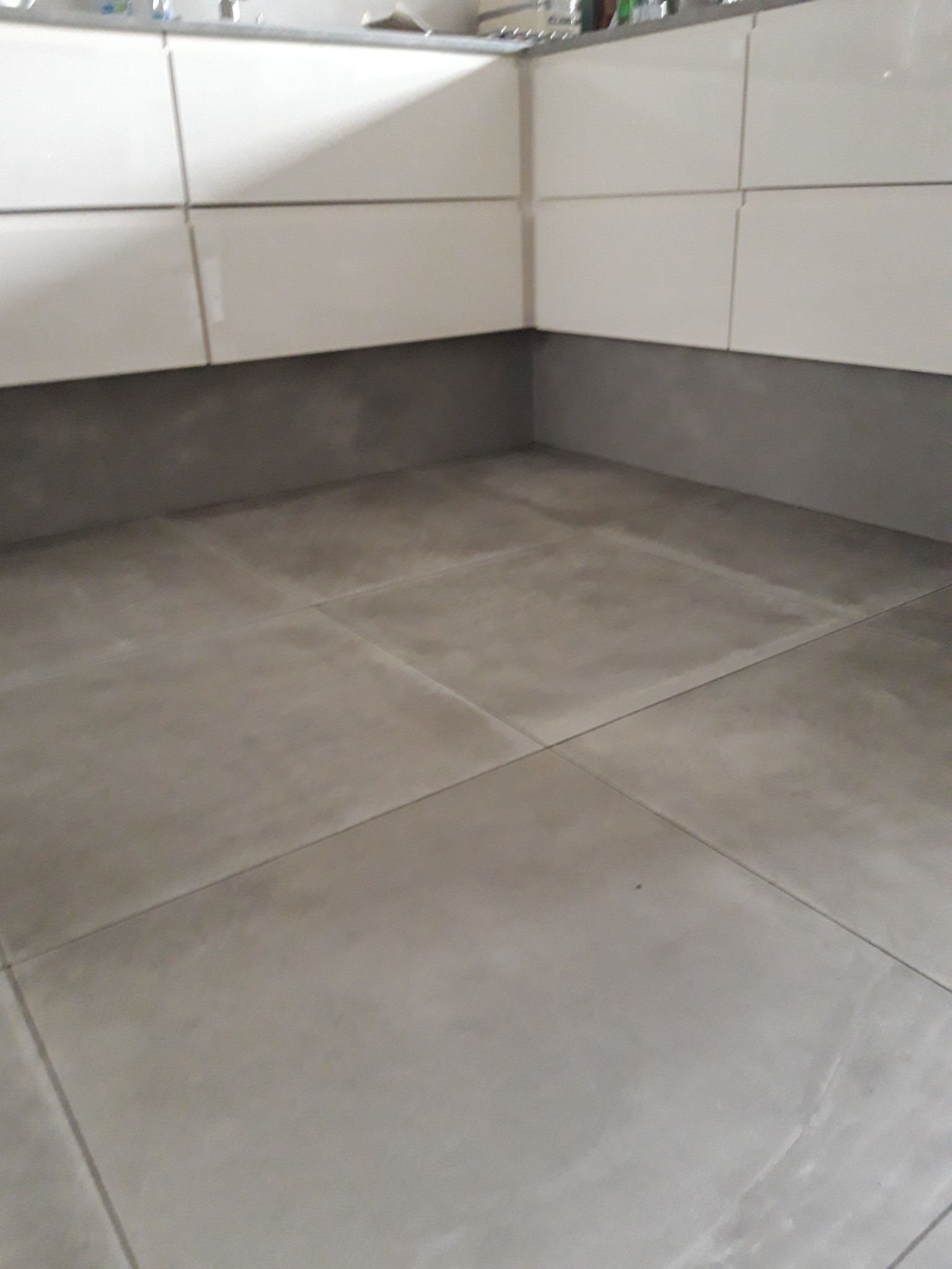 Szare, kwadratowe płytki podłogowe w łazience, z widocznymi fugami i dolną częścią białych szafek.