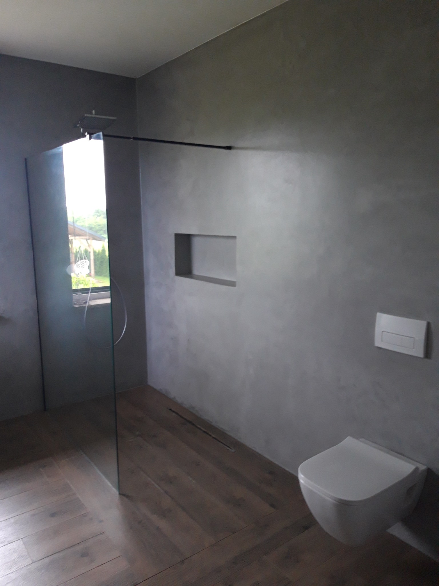 Nowoczesna łazienka z szarą ścianą wykończoną mikrocementem, kabina prysznicowa ze szklaną ścianą, drewnopodobna podłoga, wisząca toaleta i wnęka w ścianie.