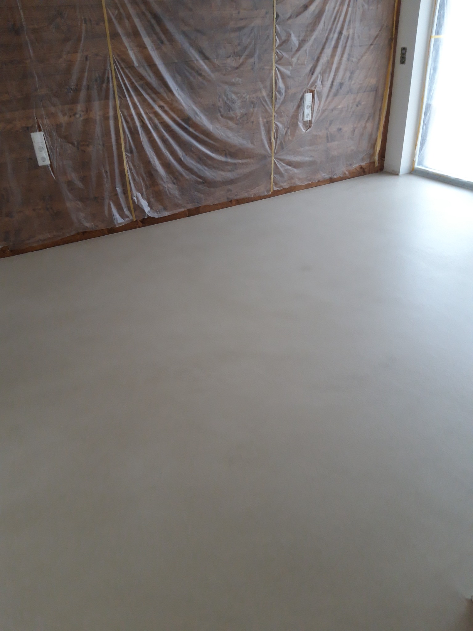 Świeżo wylana, gładka, beżowa posadzka w pomieszczeniu. Ściany zabezpieczone folią i taśmą malarską. Widoczne gniazdka elektryczne i fragment okna.