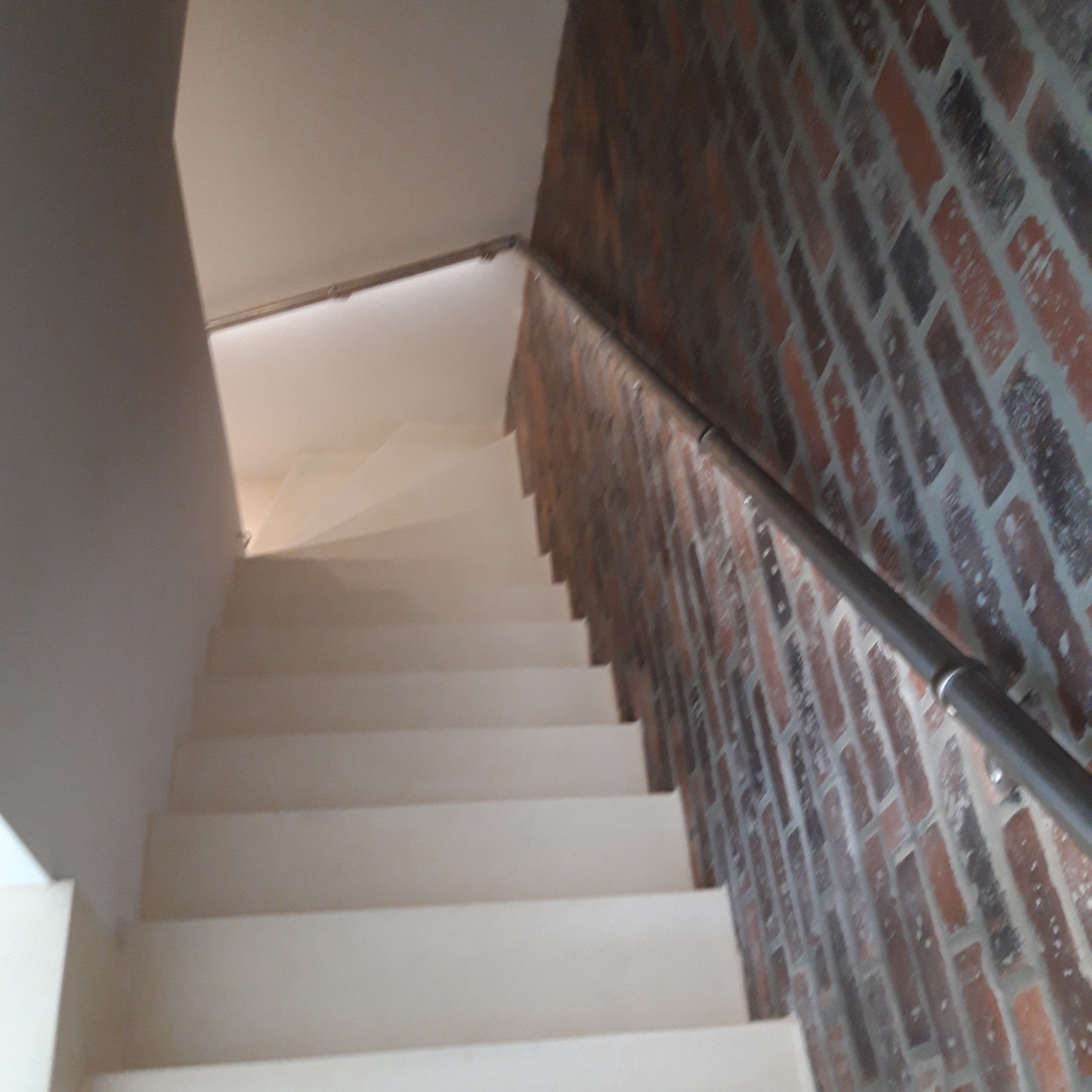 Klatka schodowa z białymi schodami i ścianą wykończoną cegłą, zabezpieczona metalową poręczą.