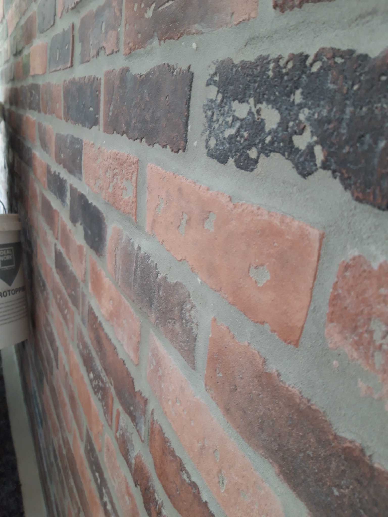 Ściana z cegły klinkierowej w odcieniach czerwieni i brązu, z widoczną fugą w kolorze szarym, ujęcie z boku z widocznym wiaderkiem z resztkami zaprawy.