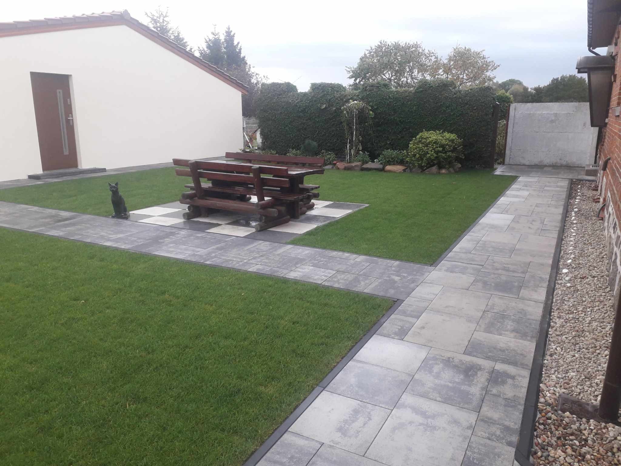 Ułożony chodnik z szarych płyt prowadzący przez trawnik, z drewnianym stołem piknikowym na szachownicy z płyt i dekoracyjną figurką kota, na tle zielonego żywopłotu i budynku z jasną elewacją.