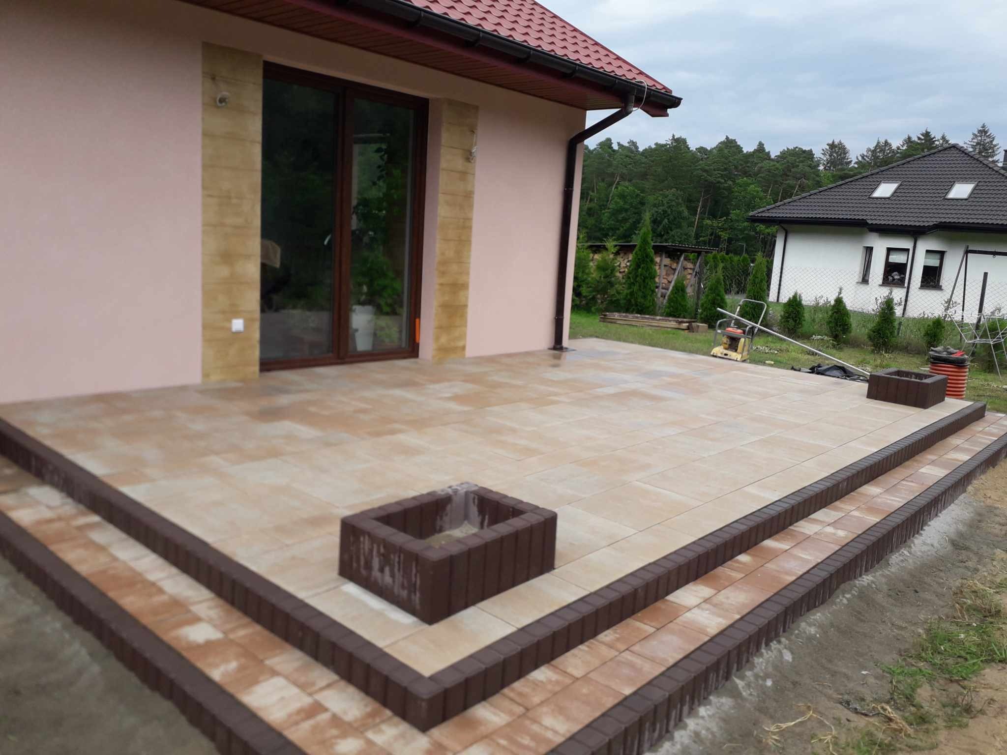 Ułożony taras z kostki brukowej w odcieniach beżu i brązu, z dwoma stopniami i elementami dekoracyjnymi w kształcie kwadratowych donic, widok z perspektywy trawnika, w tle dom z czerwoną dachówką...