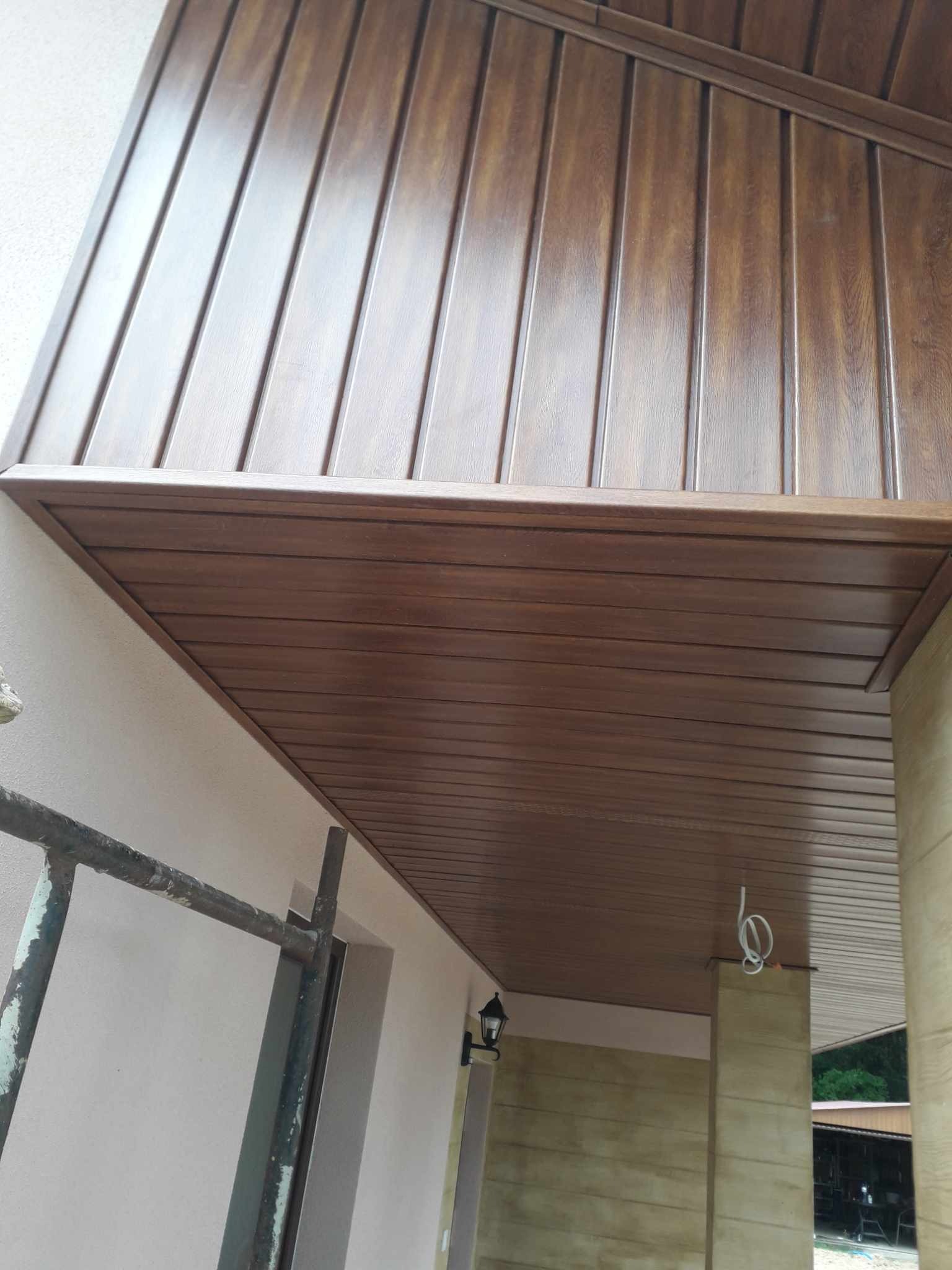 Brązowa podsufitka wykonana z paneli PCV imitujących drewno, zamontowana na zewnątrz budynku, widoczna z dołu pod kątem, z elementami rusztowania po lewej stronie.