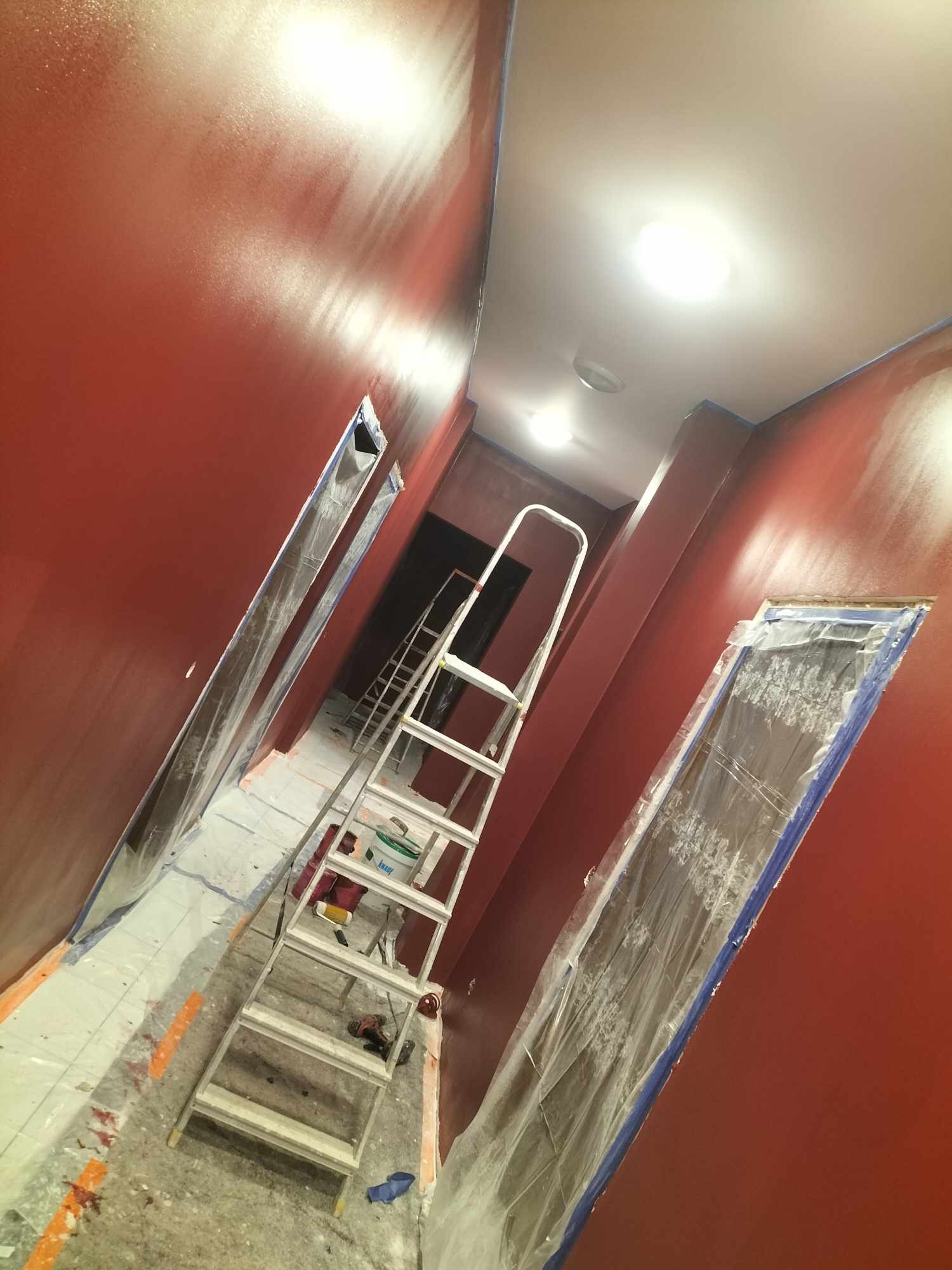 Korytarz w trakcie malowania na czerwono-brązowy kolor, zabezpieczone folią framugi drzwi, biała drabina aluminiowa na podłodze zabezpieczonej kartonem i folią malarską.