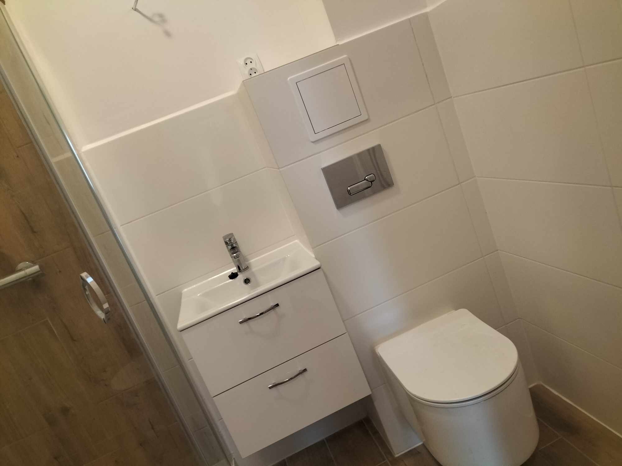 Nowoczesna łazienka z białymi płytkami, wiszącą szafką z umywalką i toaletą, widok z kabiny prysznicowej.