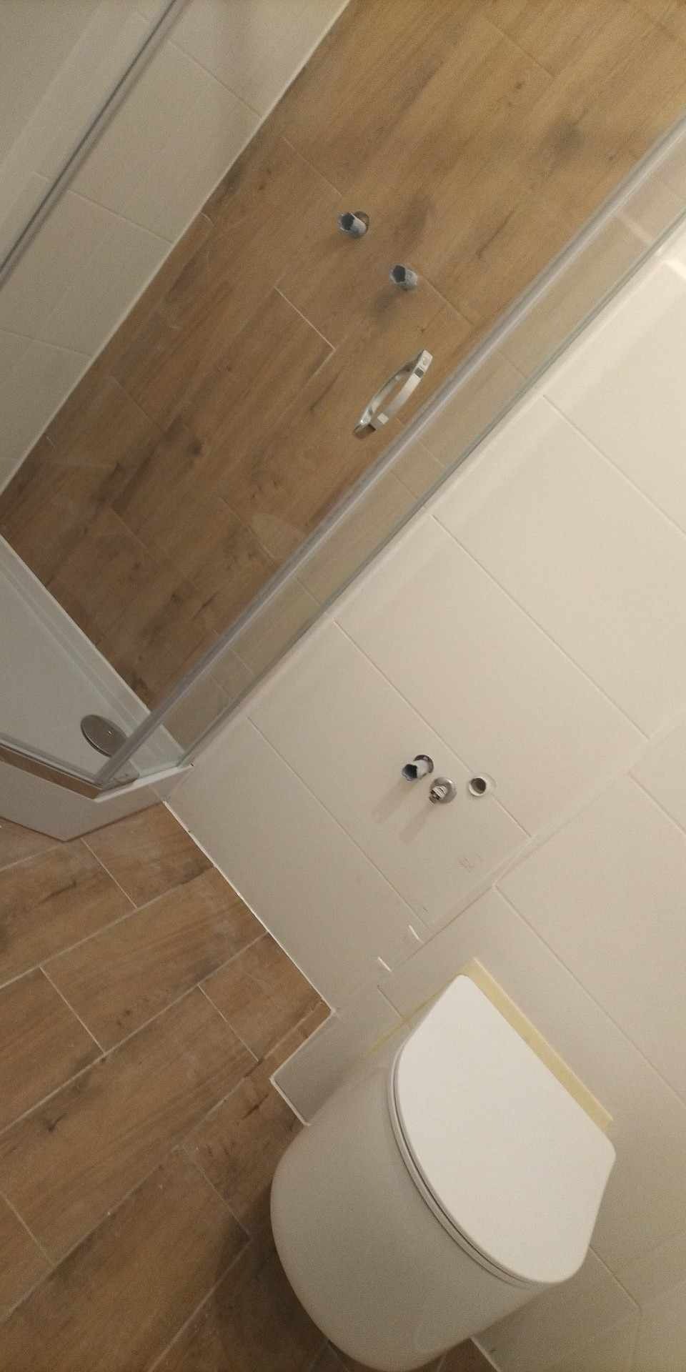 Nowoczesna łazienka w trakcie wykańczania: biała toaleta, kabina prysznicowa z przezroczystymi ściankami, płytki imitujące drewno na podłodze i ścianie prysznica, białe płytki na pozostałych...