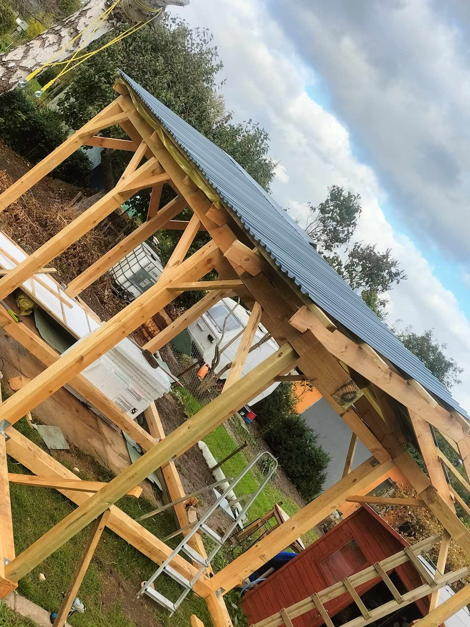 Konstrukcja drewniana dachu z blachodachówką w trakcie budowy, widok z dołu na niebo i drzewa, drabina oparta o konstrukcję.