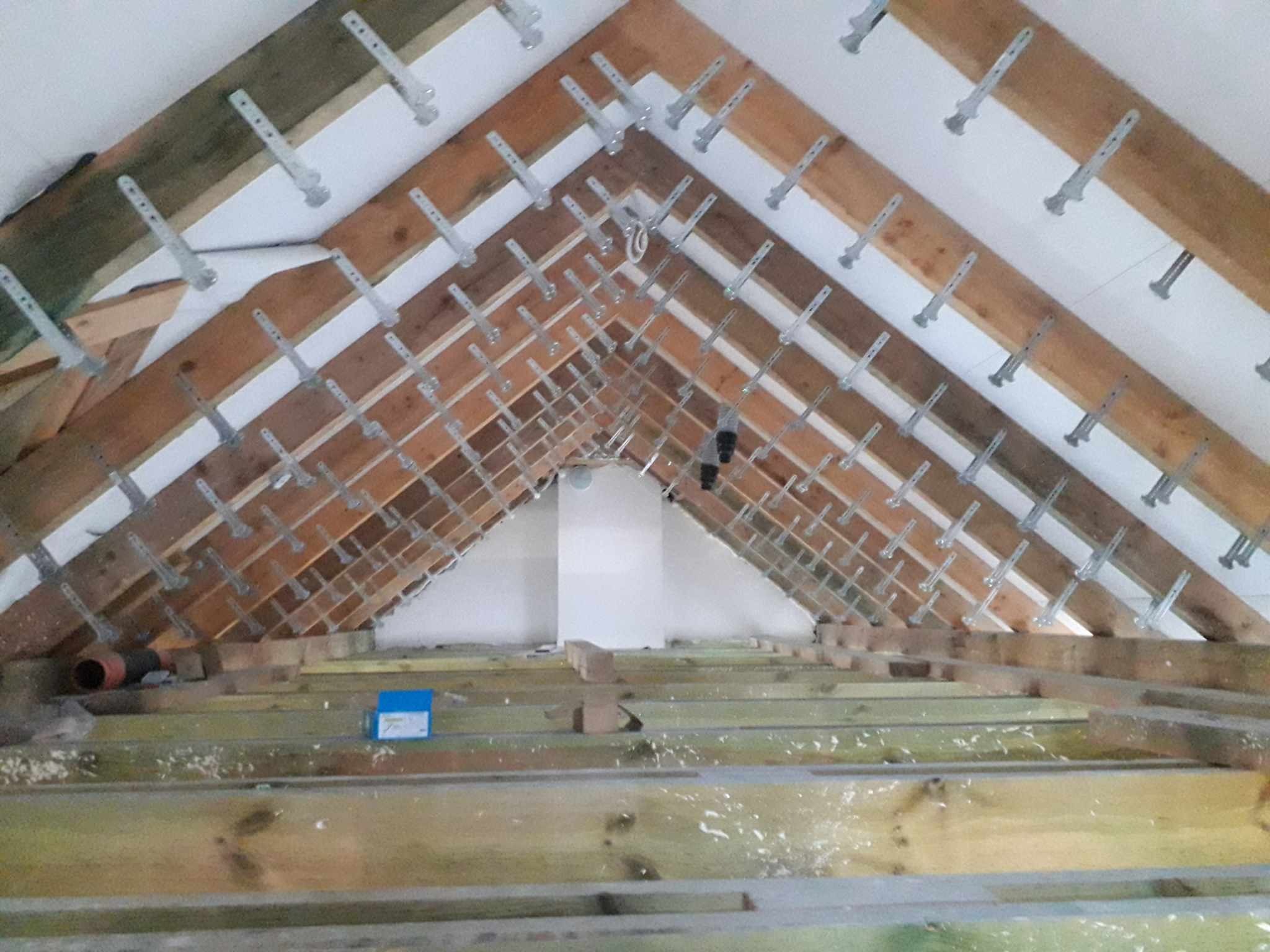 Konstrukcja drewniana poddasza z zamocowanymi metalowymi elementami mocującymi, widoczne ocieplenie między krokwiami i fragment podłogi z resztkami materiałów budowlanych.