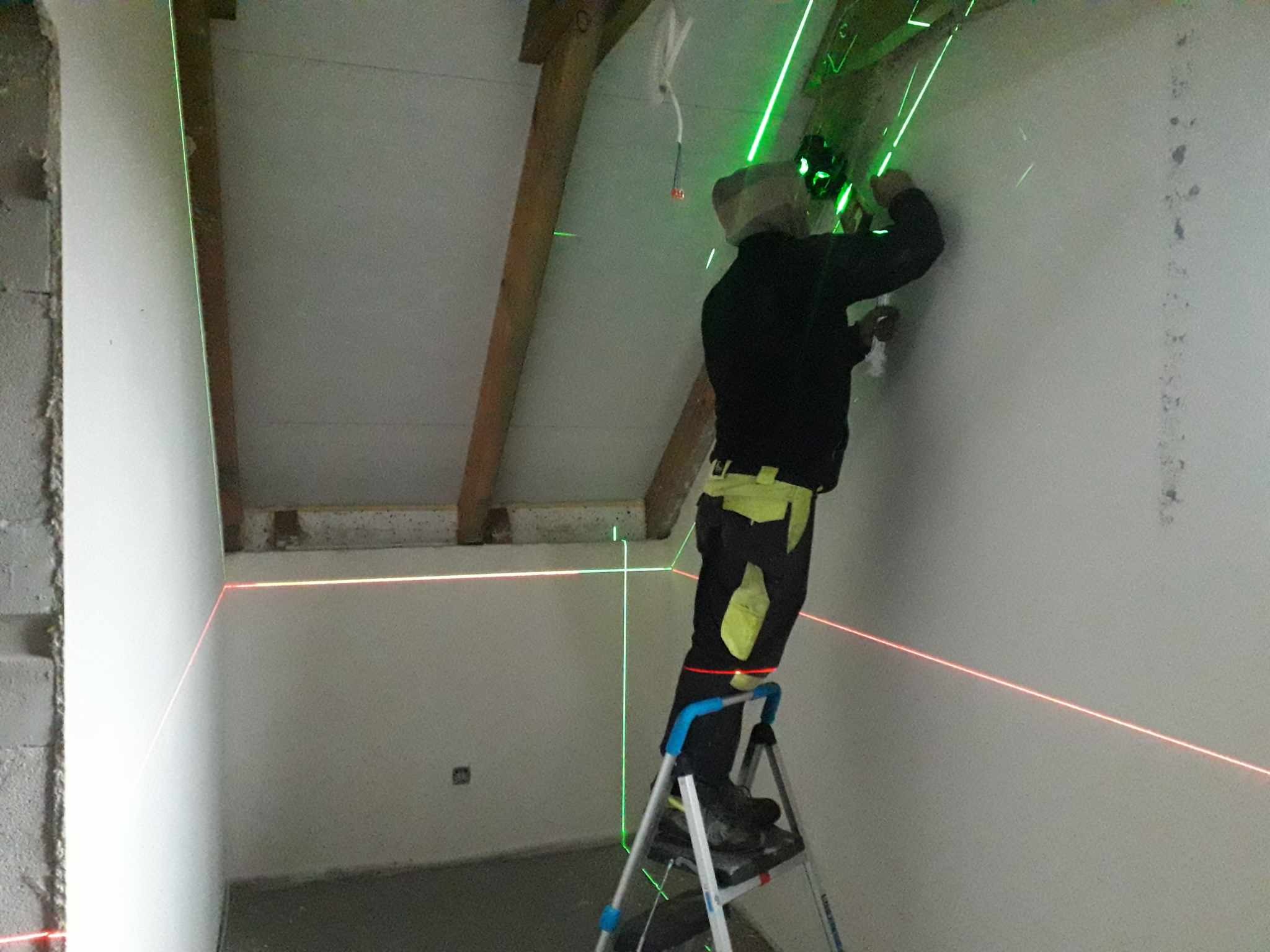 Pracownik na drabinie wyznacza poziomice laserem krzyżowym na ścianie poddasza w trakcie prac wykończeniowych.