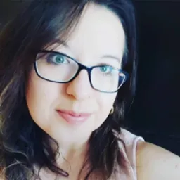 Portret uśmiechniętej kobiety z okularami w czarnych oprawkach i długimi, ciemnymi włosami, z delikatnym makijażem, na ciemnym tle.