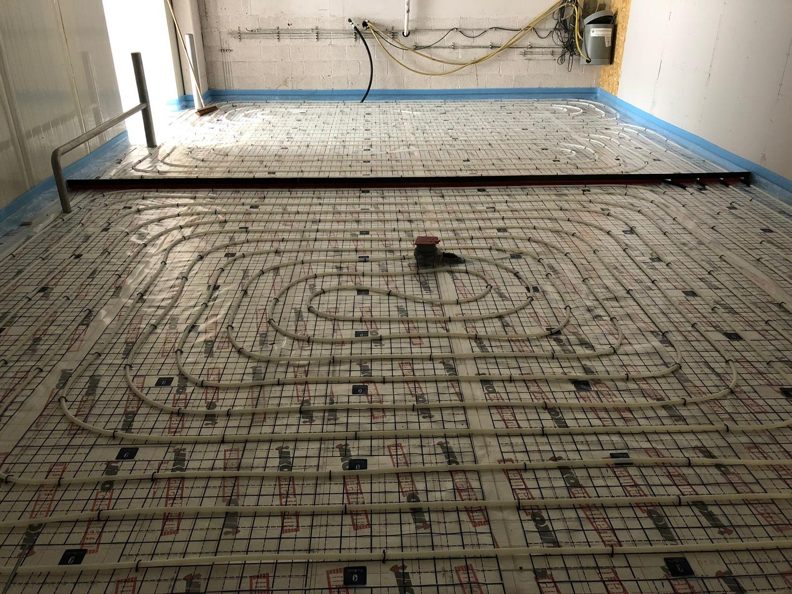 Instalacja ogrzewania podłogowego: widok z góry na rozłożone rury grzewcze w systemie pętli, zamocowane na siatce z folią izolacyjną w surowym pomieszczeniu z betonową ścianą.