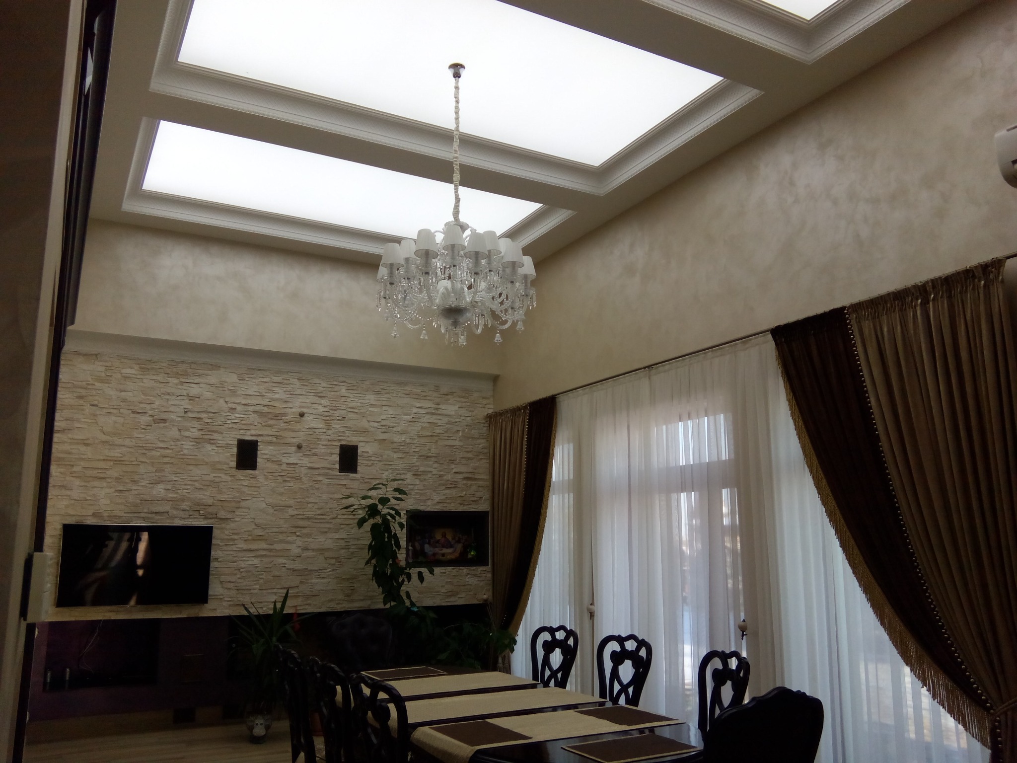 Eleganckie oświetlenie sufitowe w jadalni z żyrandolem, podwieszany sufit z panelami LED. Stonowane barwy ścian i dekoracyjne zasłony. Widok na stół jadalny i krzesła.