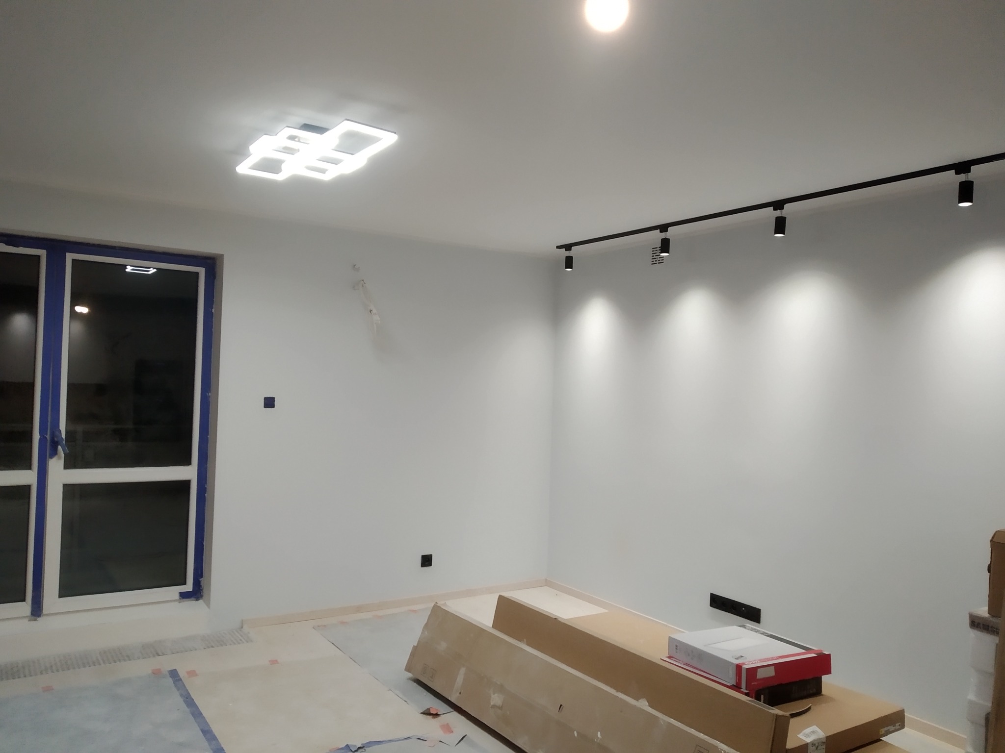 Nowoczesne oświetlenie sufitowe LED: geometryczny plafon i szyna z reflektorami w jasnym wnętrzu. Okno z niebieskimi ramami i kartony na podłodze.