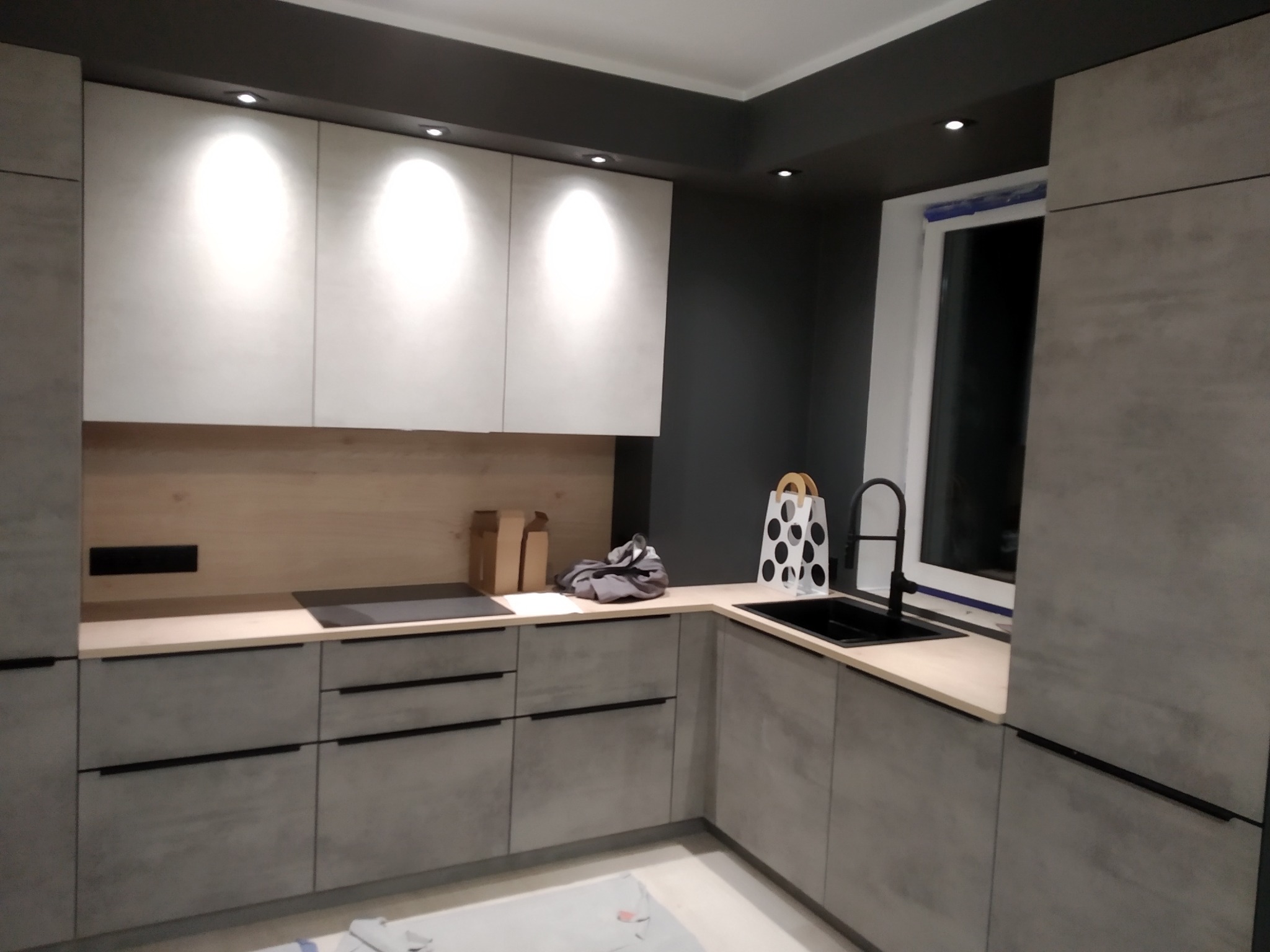 Nowoczesna kuchnia w szarościach z białymi szafkami górnymi i oświetleniem punktowym. Na blacie widoczna płyta indukcyjna.