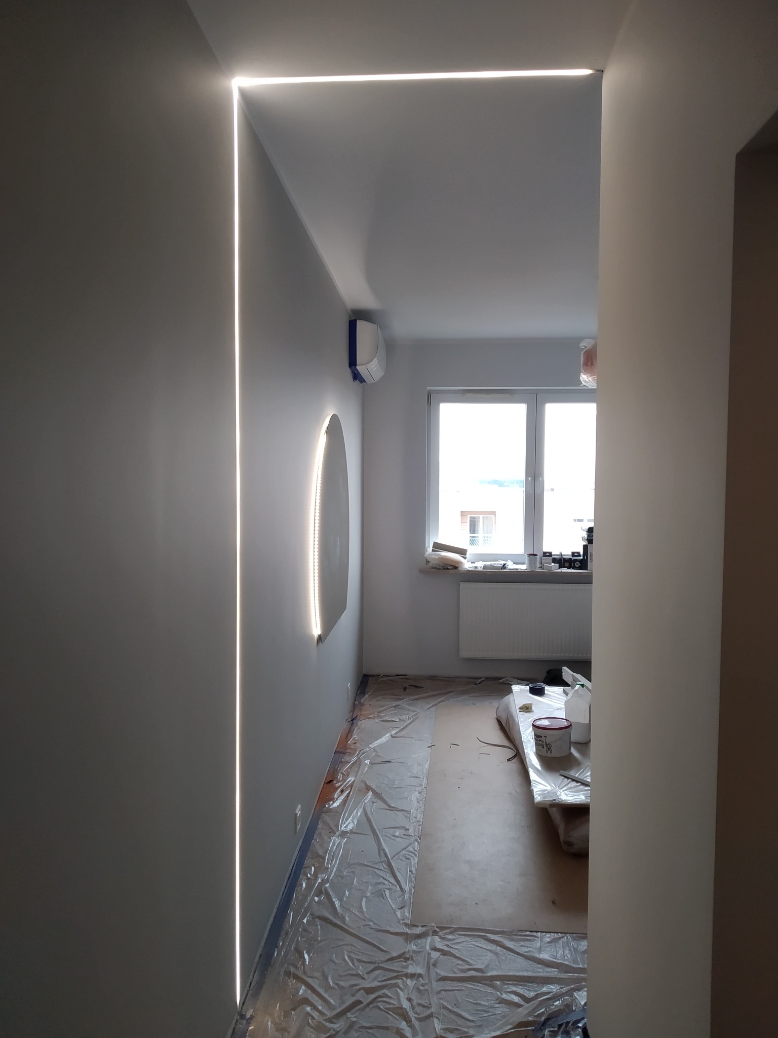 Nowoczesne oświetlenie LED w korytarzu z ukrytą listwą świetlną w kształcie litery L, oraz dekoracyjna lampa ścienna. Widoczne przygotowania do malowania ścian.