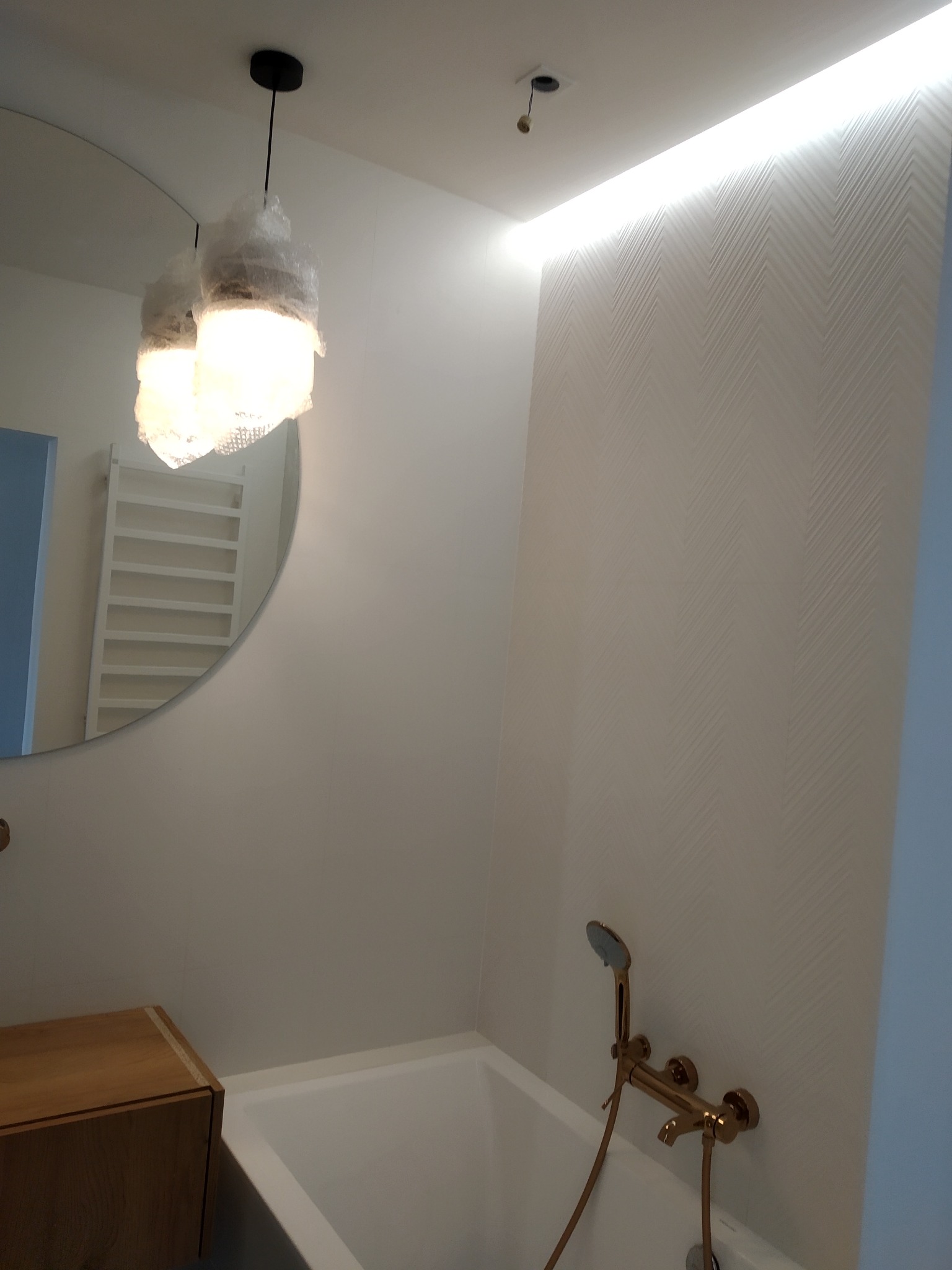 Nowoczesna łazienka z podświetleniem LED na suficie i lampami wiszącymi w folii ochronnej. Złota armatura, wzorzysta ściana i okrągłe lustro.