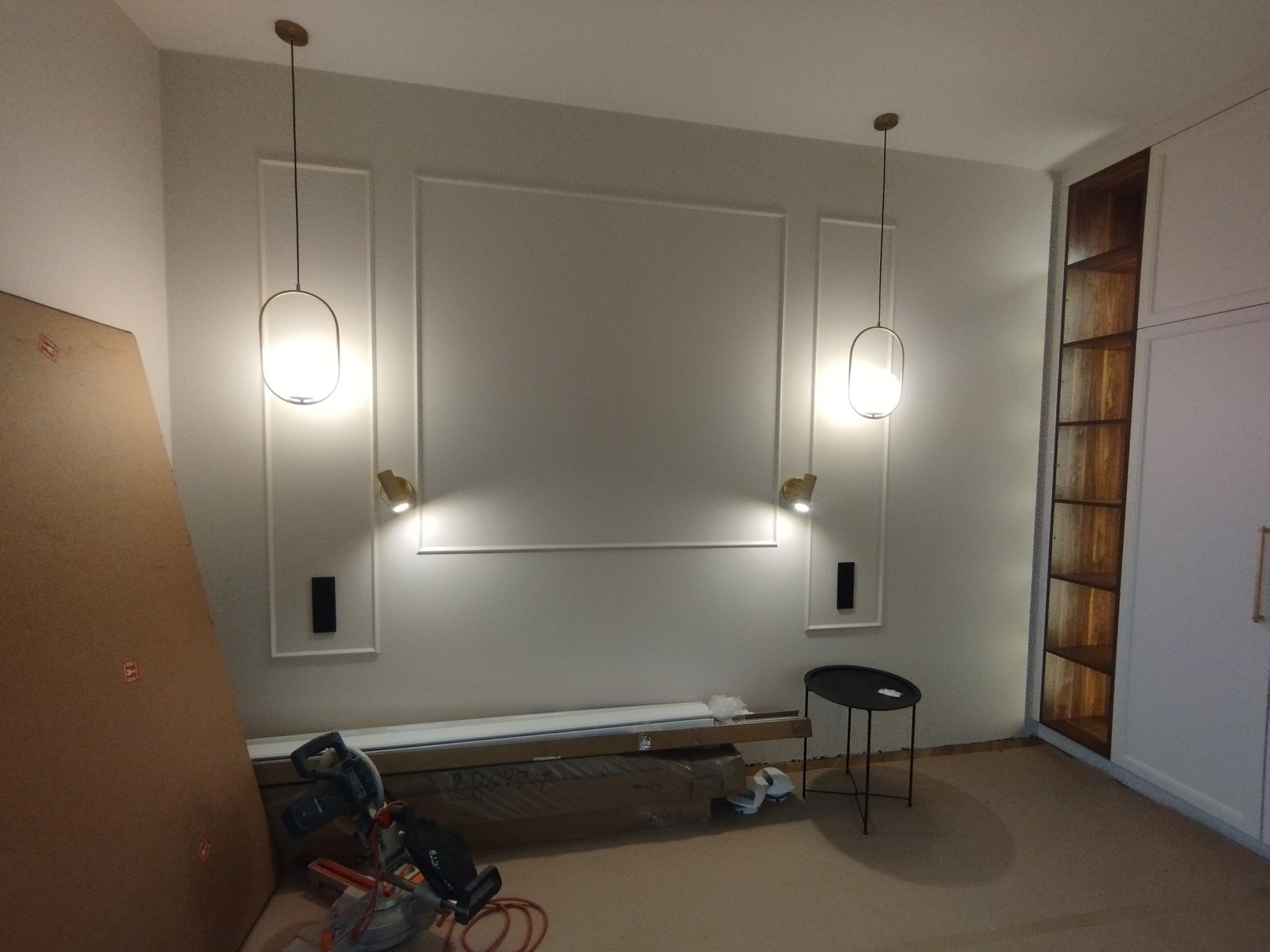 Montaż oświetlenia: Dwa złote kinkiety i lampy wiszące w pokoju z dekoracyjnymi listwami ściennymi. Na podłodze narzędzia, w tle regał z drewnianymi półkami.