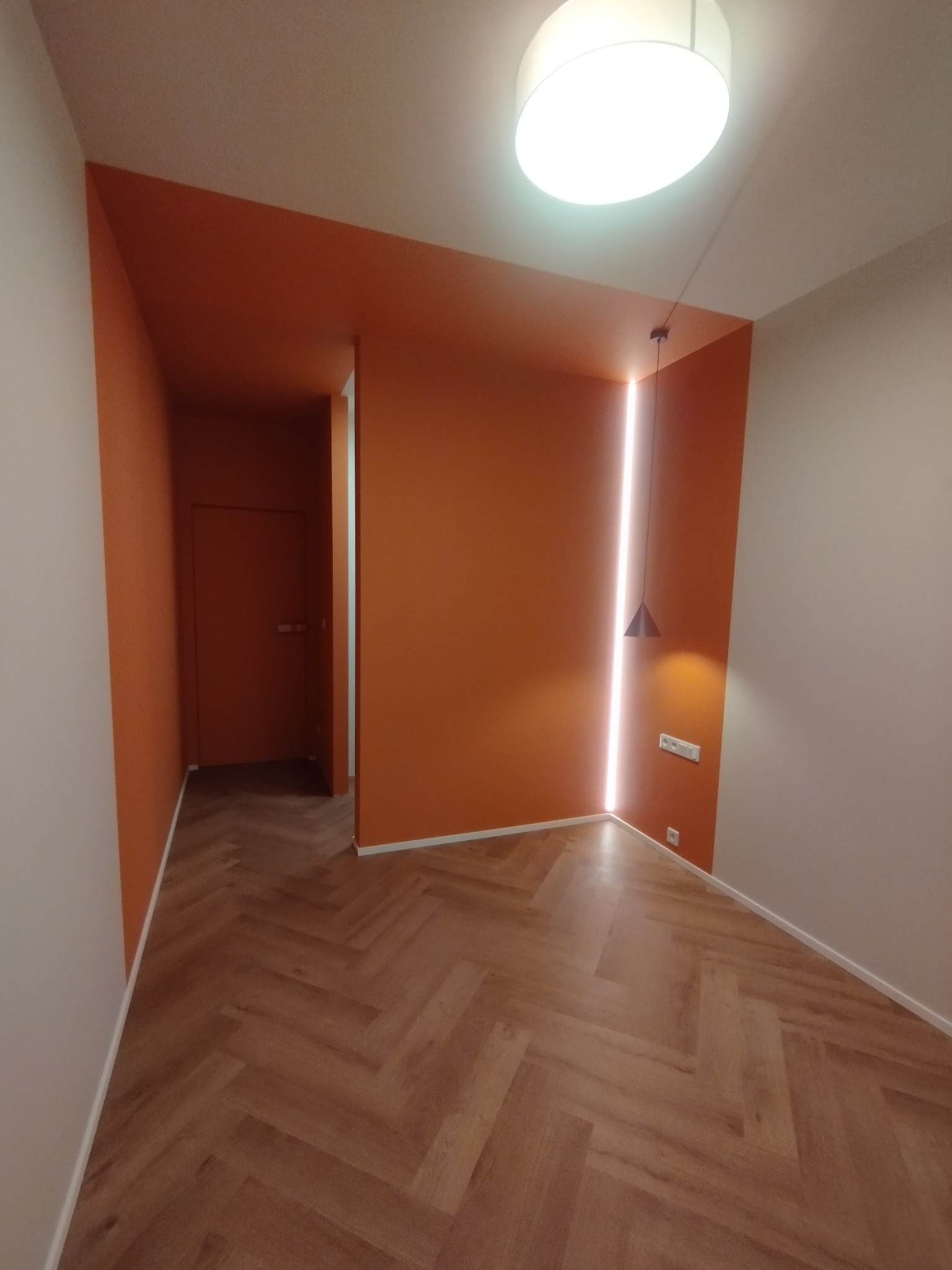 Pomarańczowe ściany z oświetleniem LED w wąskiej szczelinie, lampa wisząca, parkiet w jodełkę. Widok na korytarz z drzwiami. Nowoczesne wykończenie wnętrz.