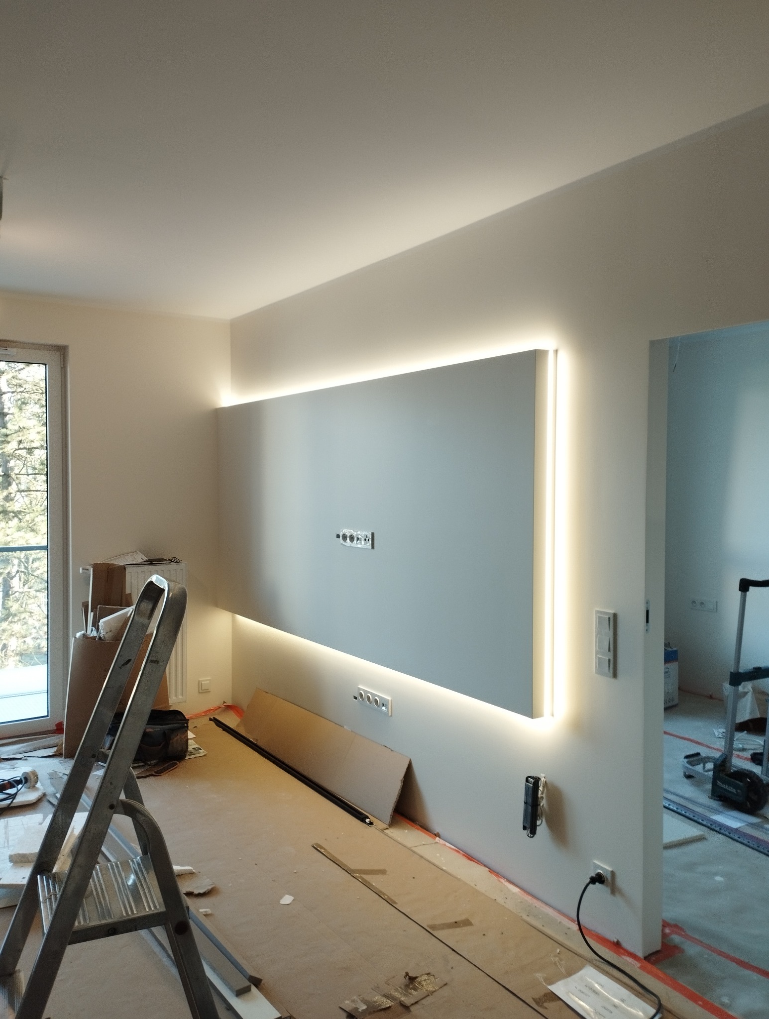 Nowoczesna ściana z podświetleniem LED w trakcie montażu. Widoczne gniazdka, drabina i narzędzia. Etap prac wykończeniowych w jasnym wnętrzu.