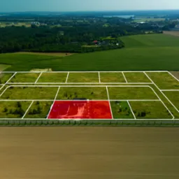 Widok z lotu ptaka na działki budowlane z zaznaczoną na czerwono jedną z nich, otoczone polami i lasami.